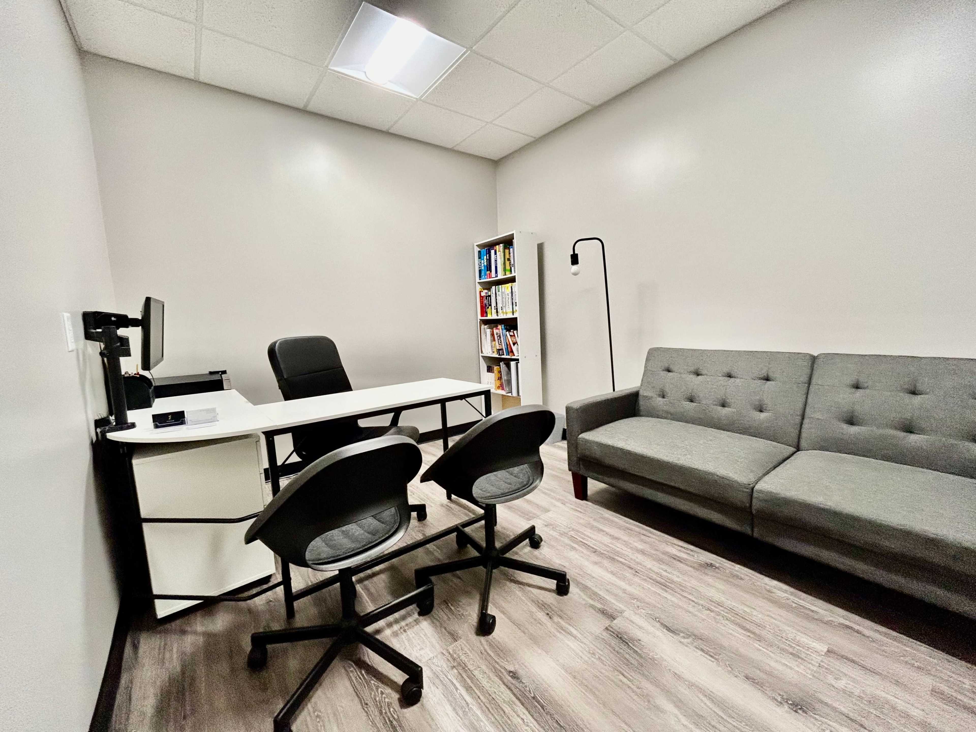 A small office space featuring a desk with a computer, two chairs, a couch, and a bookcase filled with books.