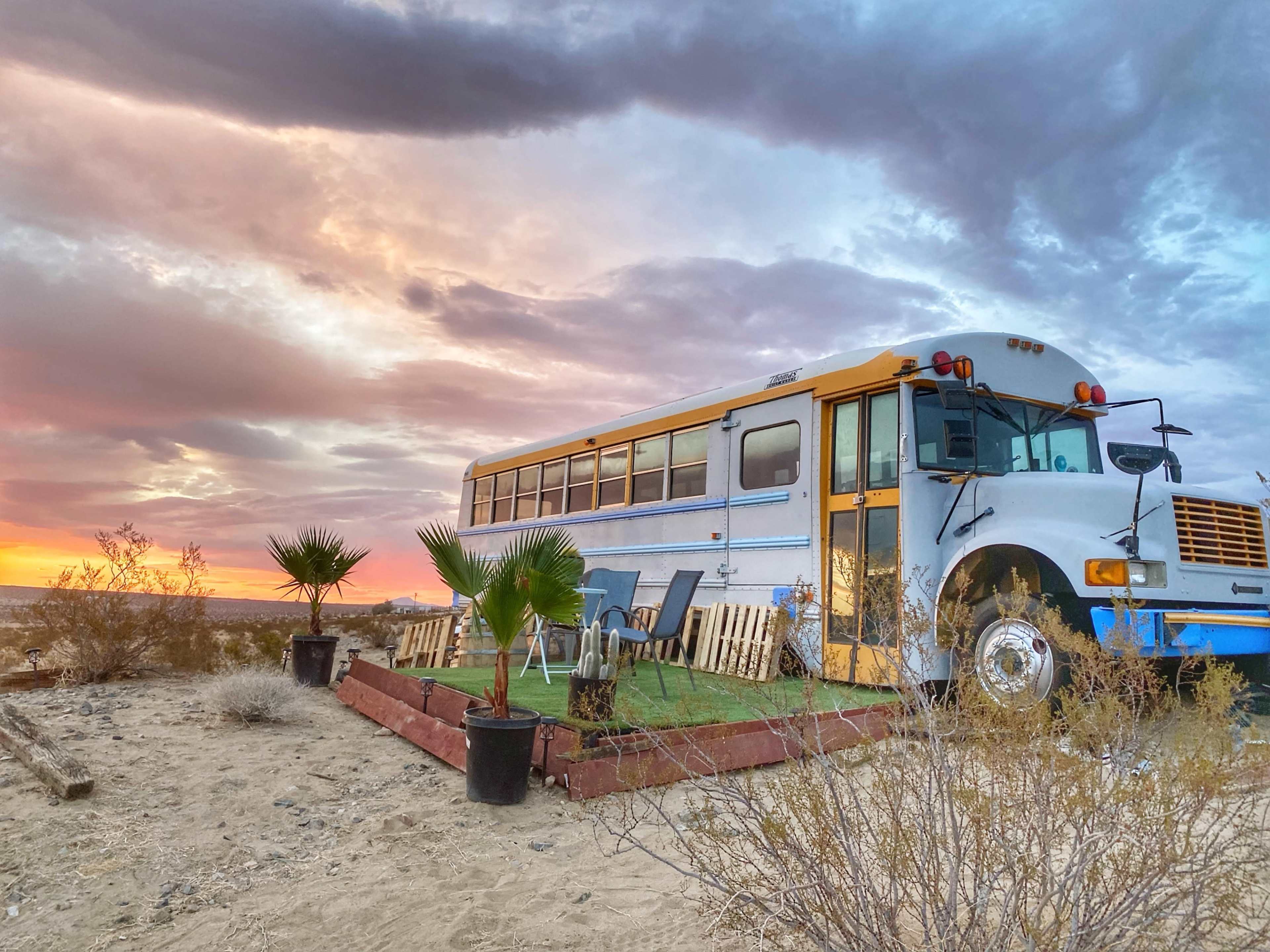 A converted school bus sits on a sandy lot with outdoor seating and desert vegetation against a colorful sunset sky.