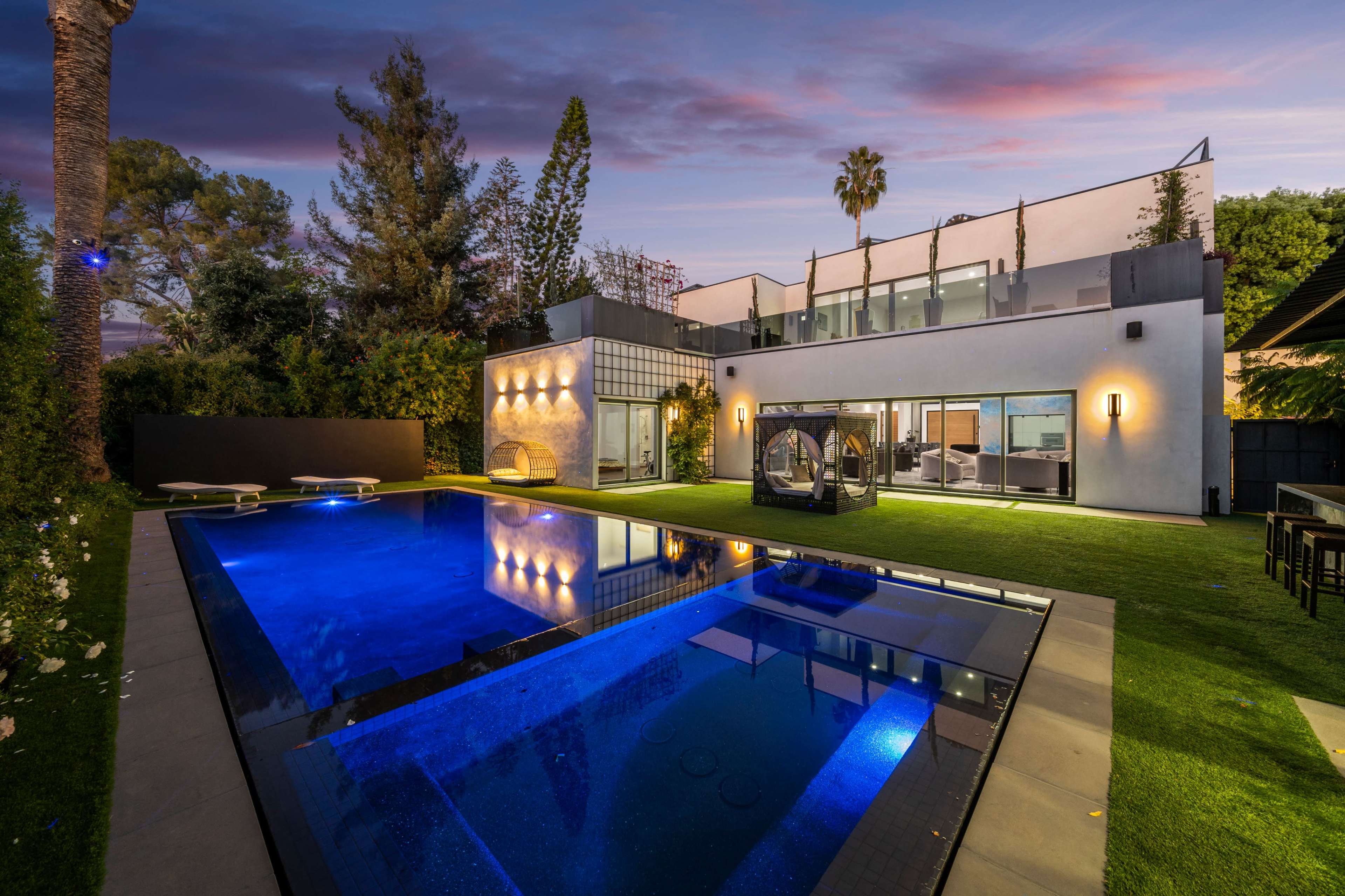 A modern house with large windows overlooks a blue swimming pool surrounded by a manicured lawn and palm trees at dusk.