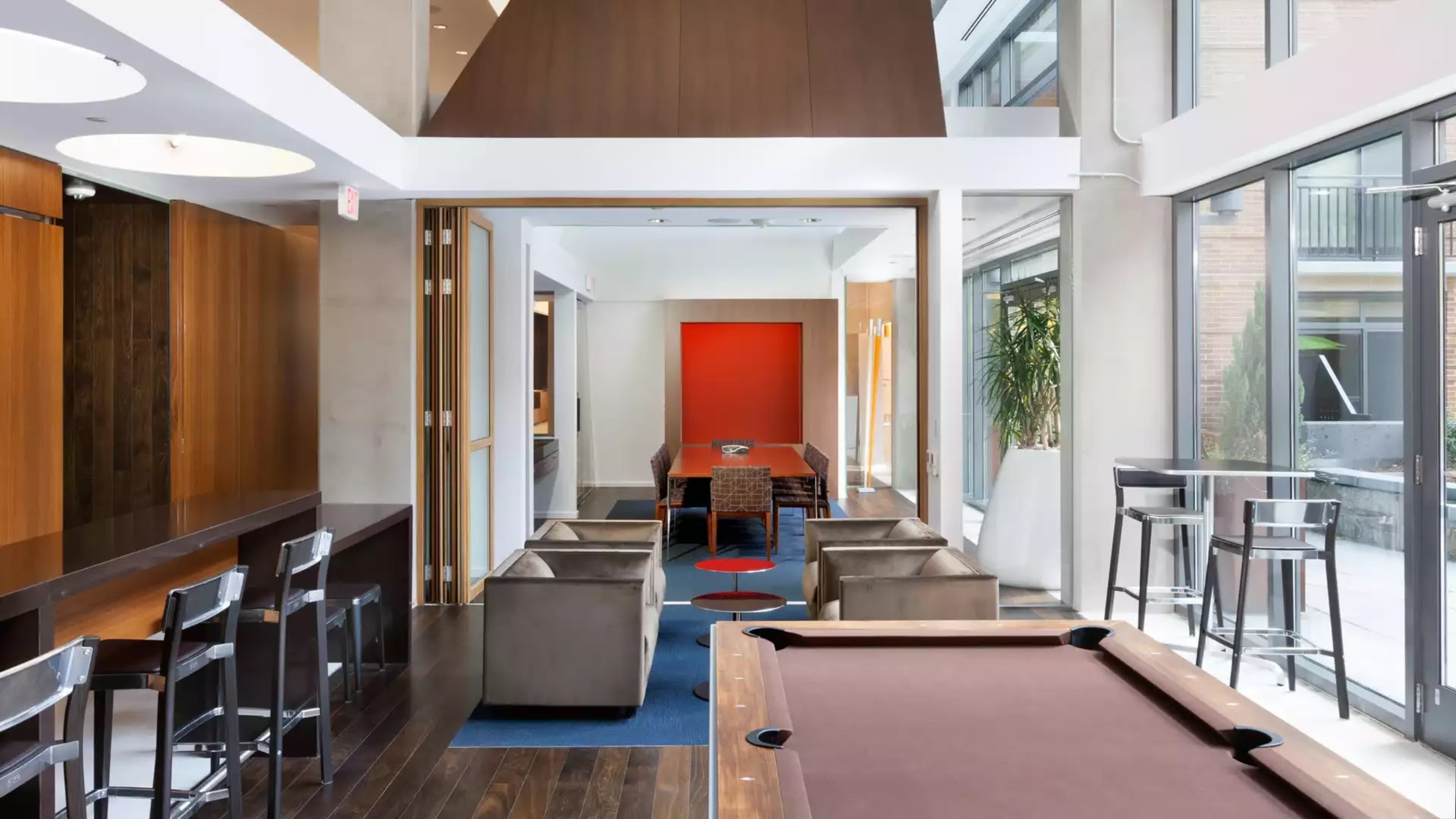 A modern open-concept lounge area featuring a billiards table, seating arrangements, and a dining space with large windows allowing natural light.