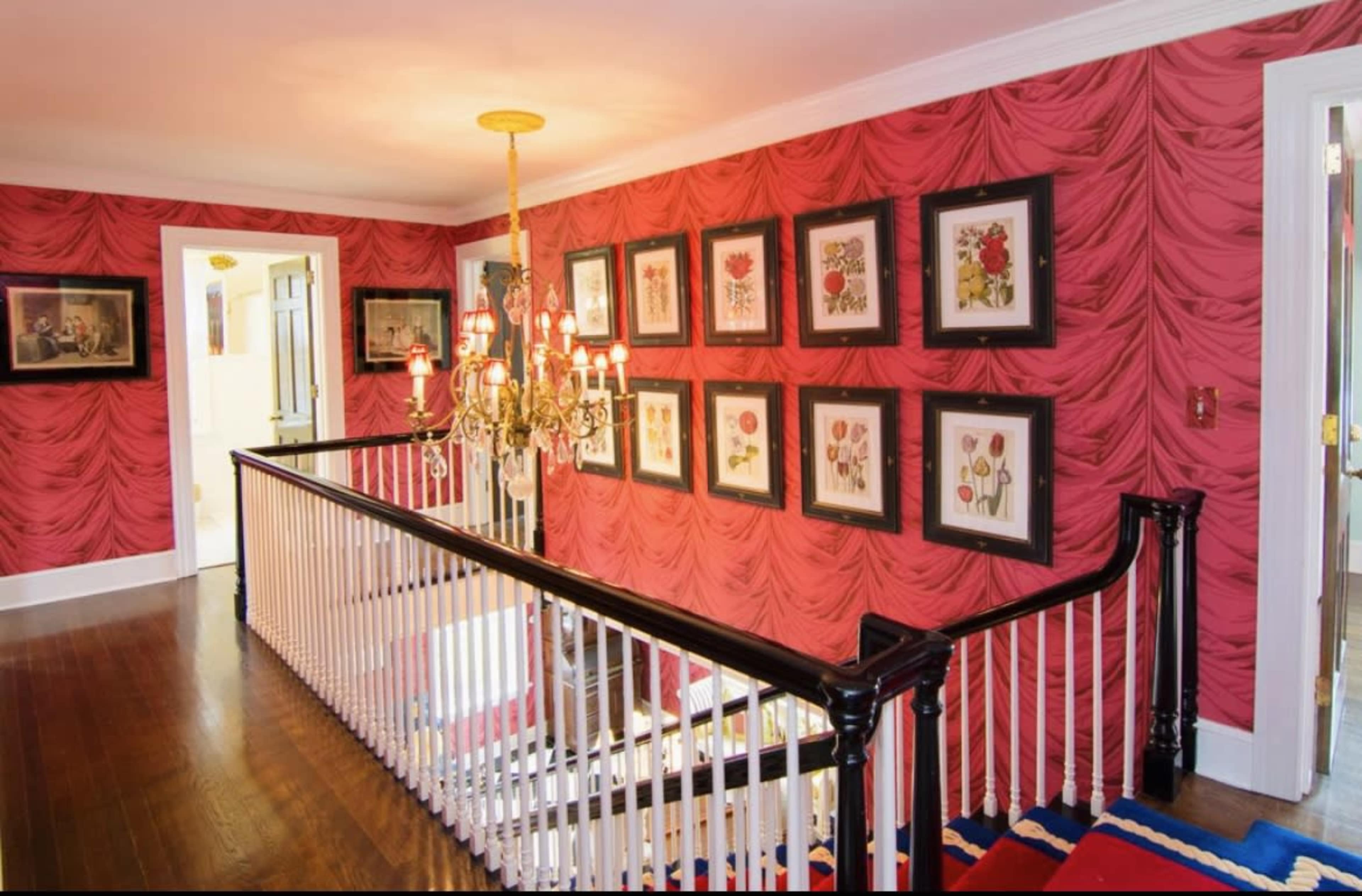 A hallway featuring a decorative red wallpaper, framed artwork on the walls, and a chandelier above a staircase.