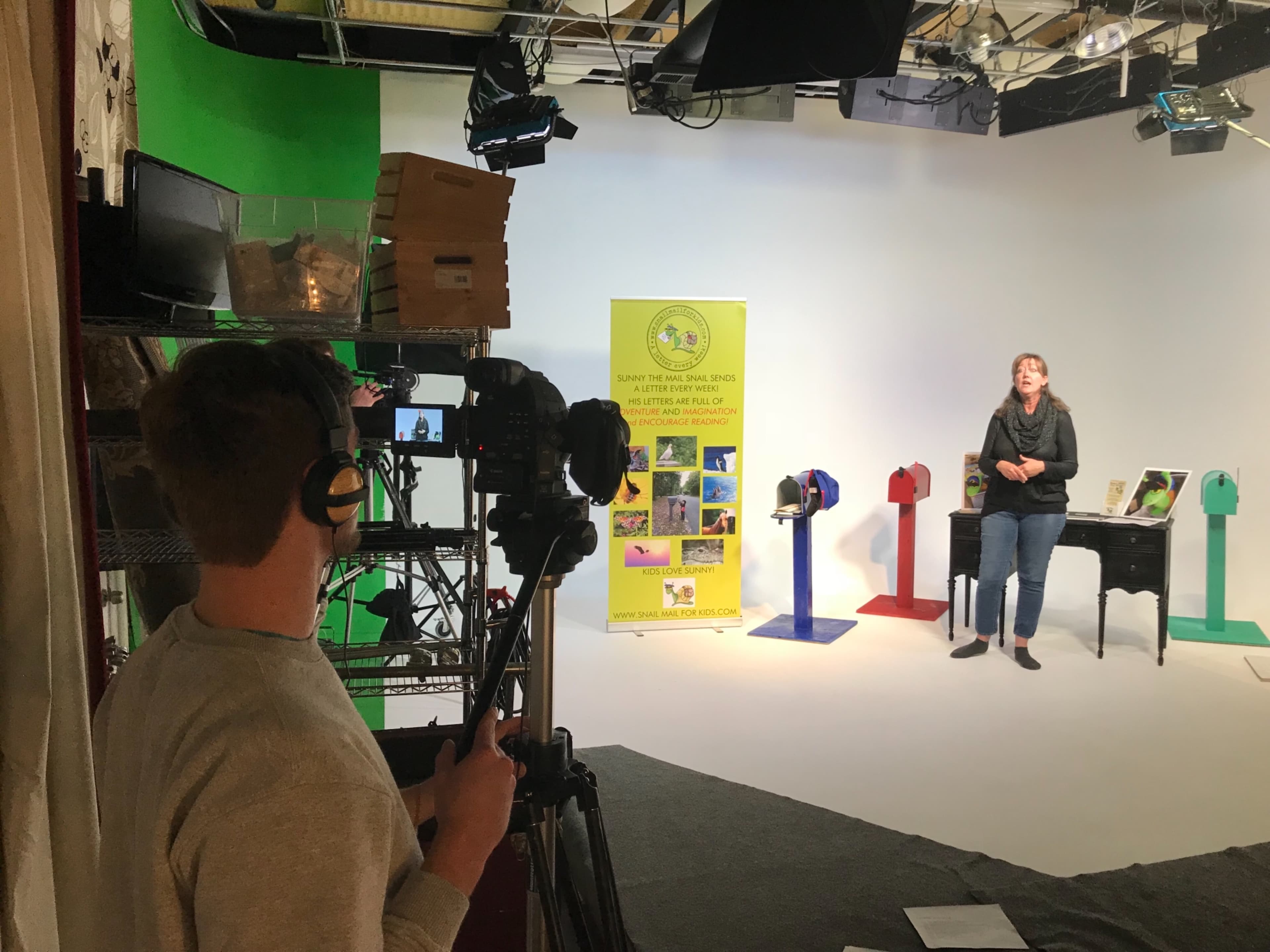A person operates a camera while recording a presenter standing next to colorful display mailboxes in a studio setting.