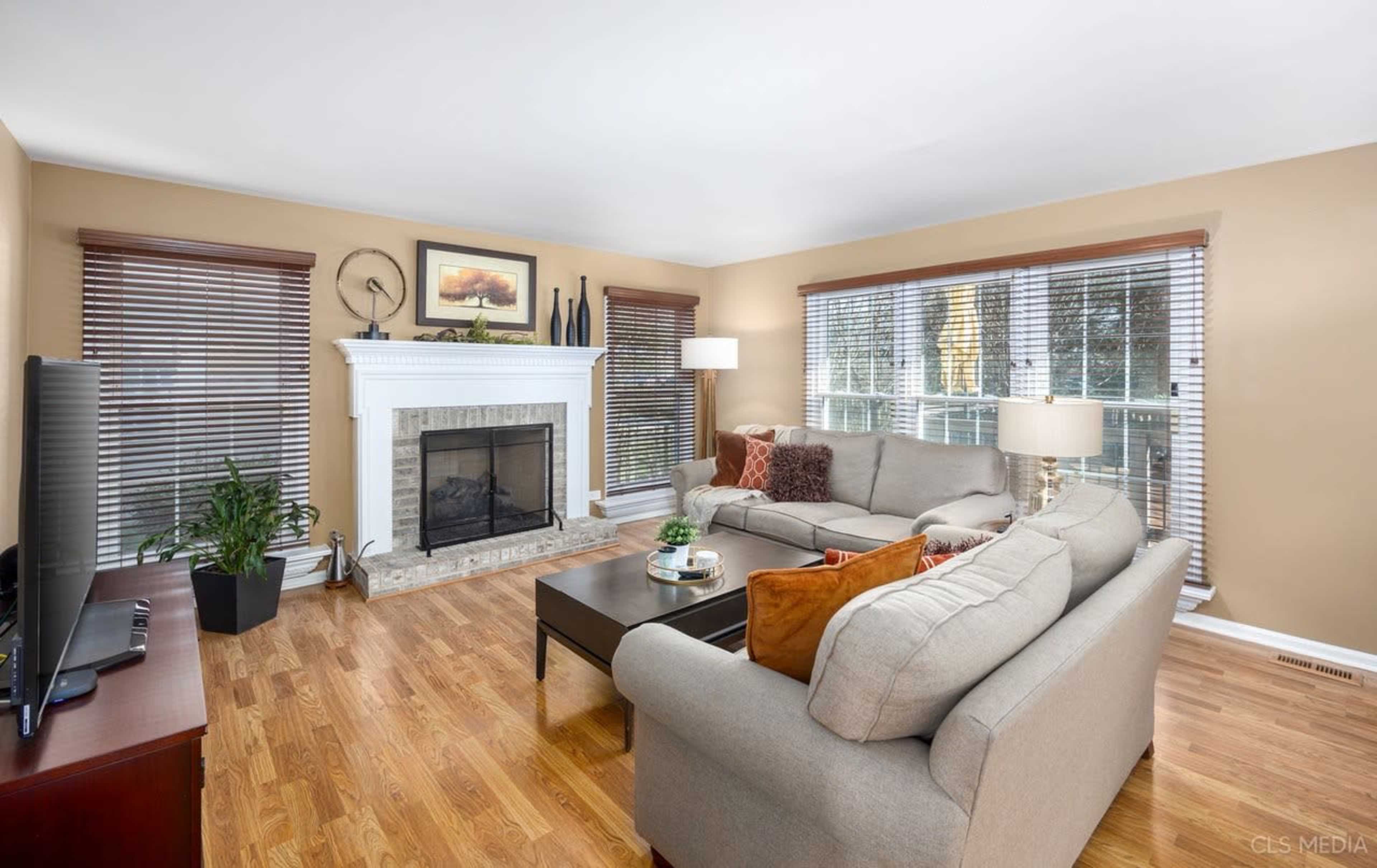 The image shows a living room with a fireplace, large windows with blinds, and a sectional sofa arranged around a coffee table.