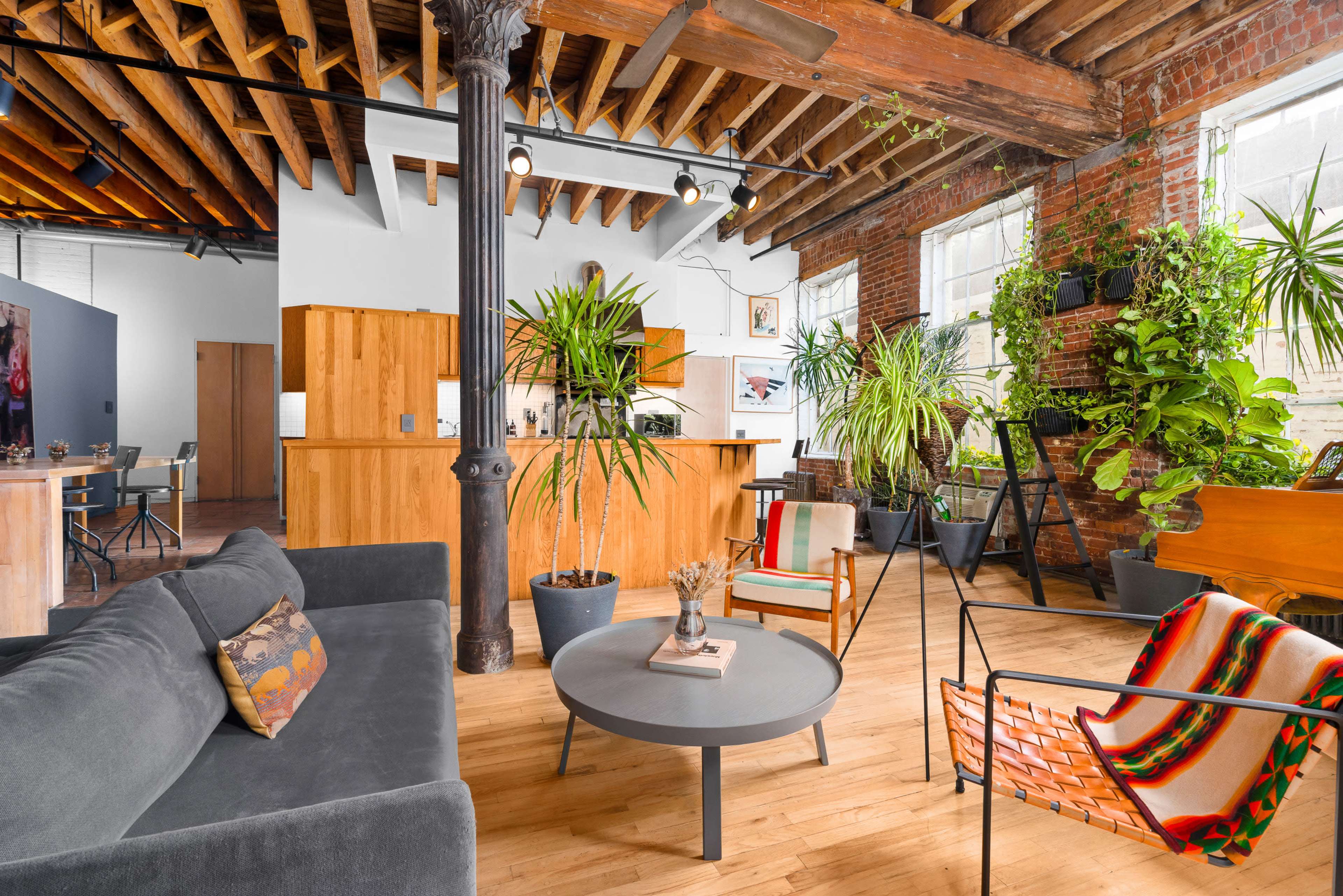 A modern living space featuring a gray sofa, a circular coffee table, and several potted plants, with exposed wooden beams and brick walls.