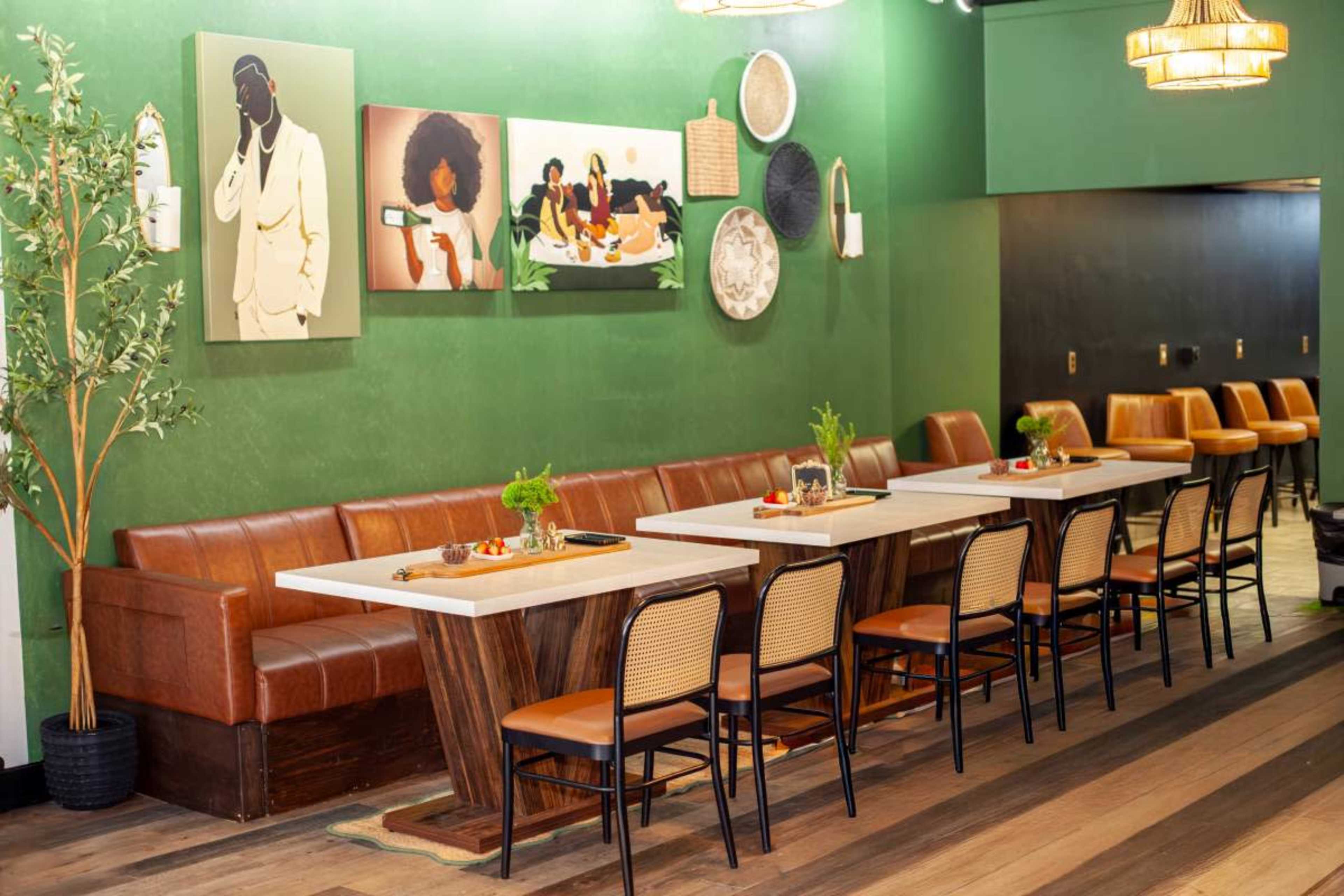 The image shows a modern restaurant interior with a green wall, several artworks, a leather bench, and tables set for dining.