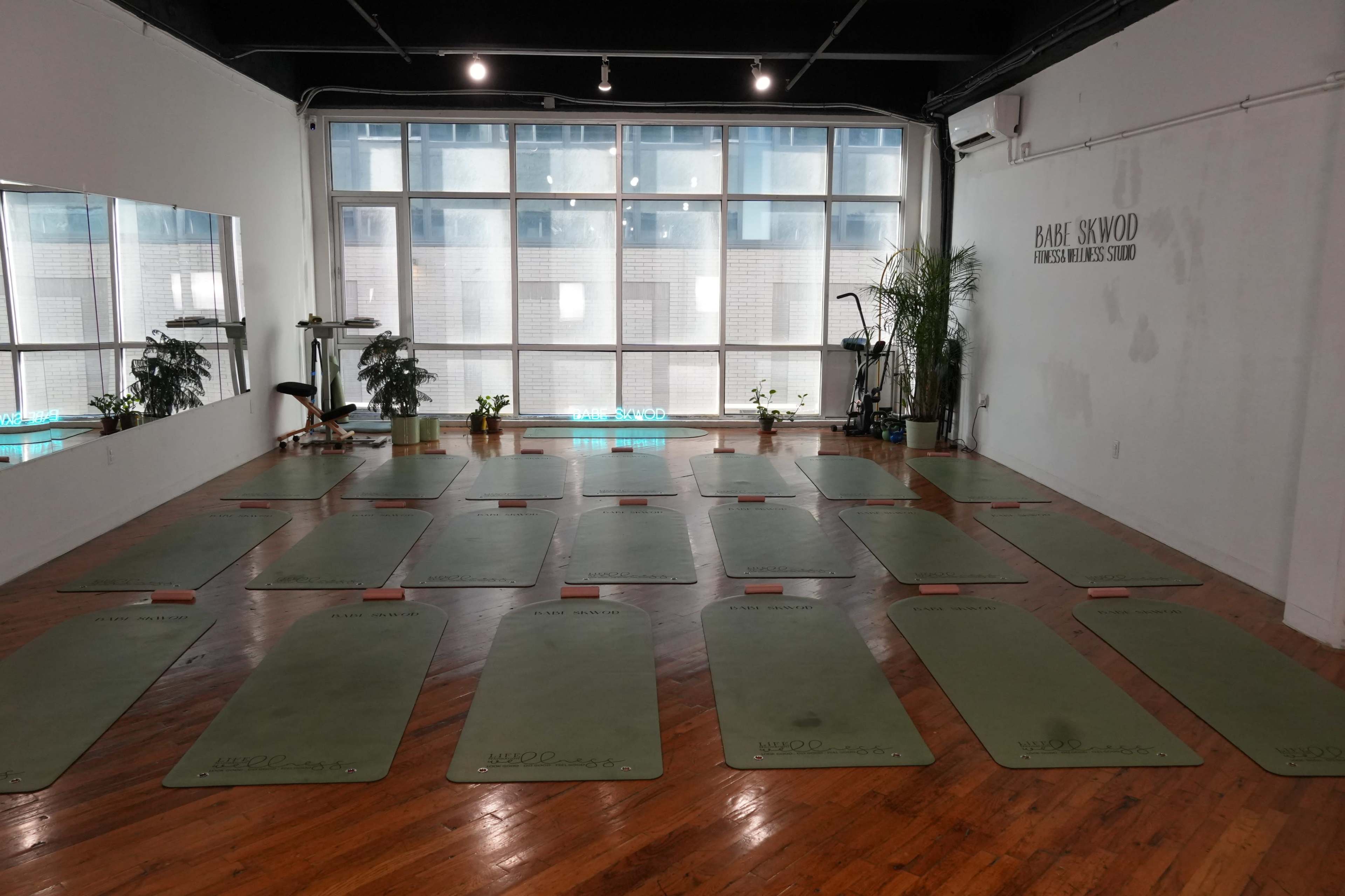 The image shows a spacious yoga studio with neatly arranged mats on a wooden floor and large windows letting in natural light.