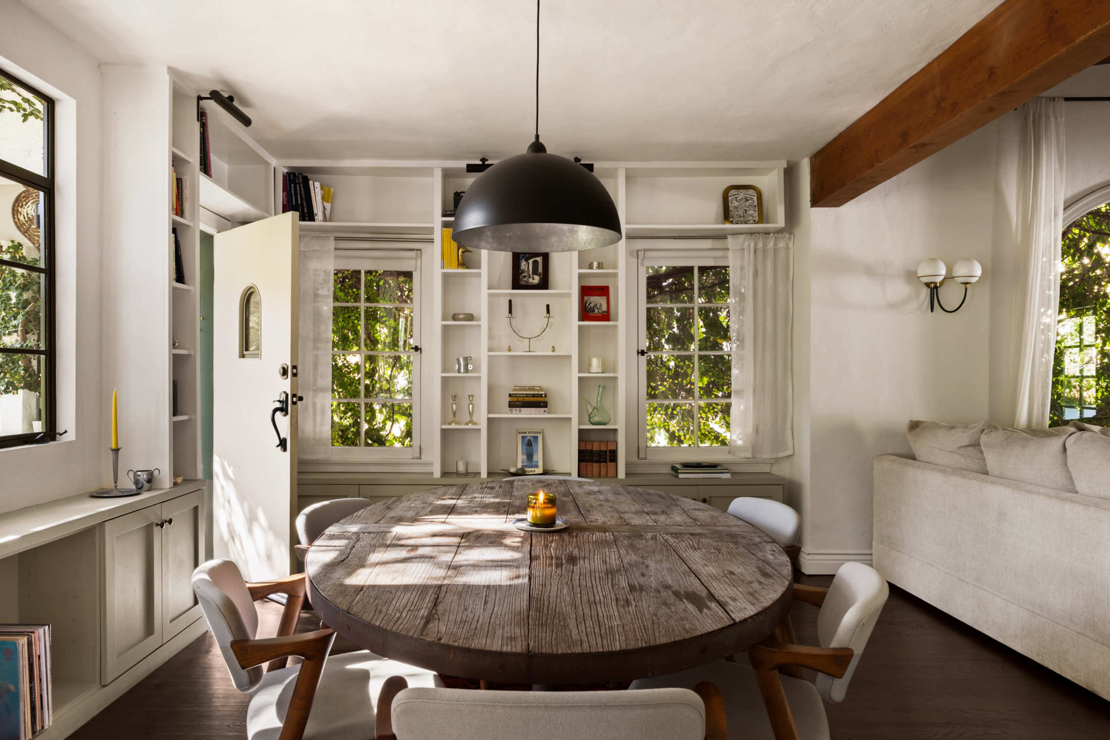 The image shows a dining area with a round wooden table surrounded by upholstered chairs, a bookcase filled with items, and windows that provide natural light.