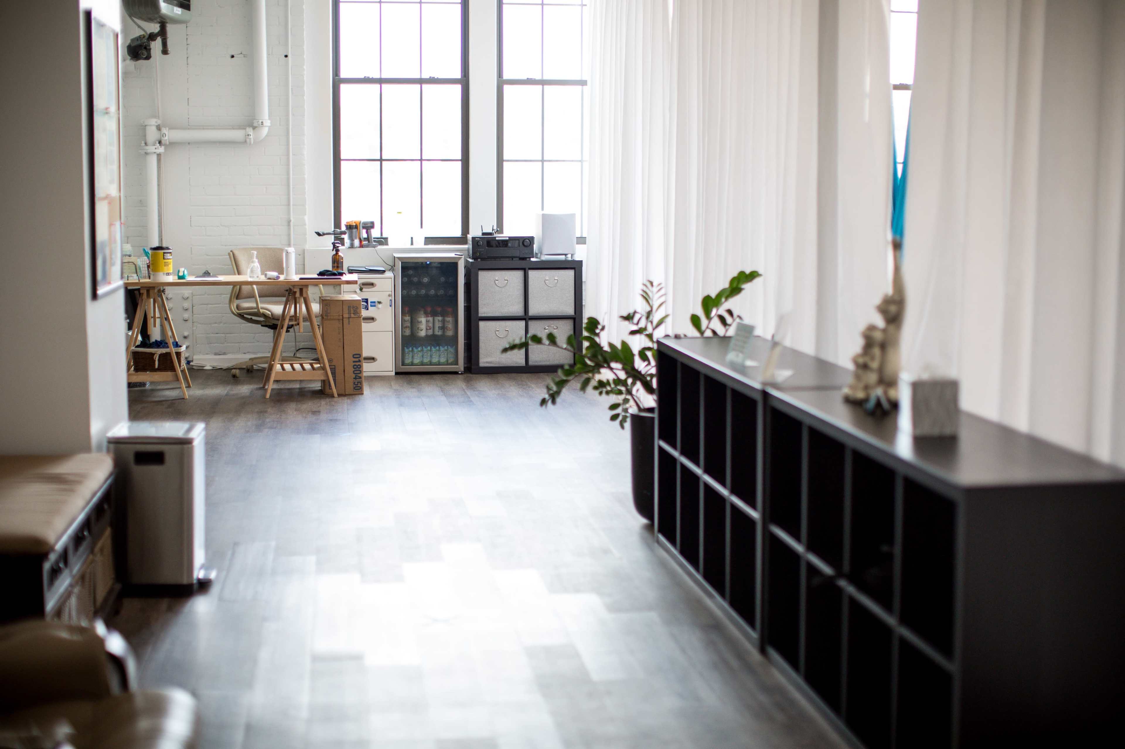 A spacious, minimalist interior with a desk and office equipment on one side and a bookshelf on the other, separated by a corridor with large windows allowing natural light.