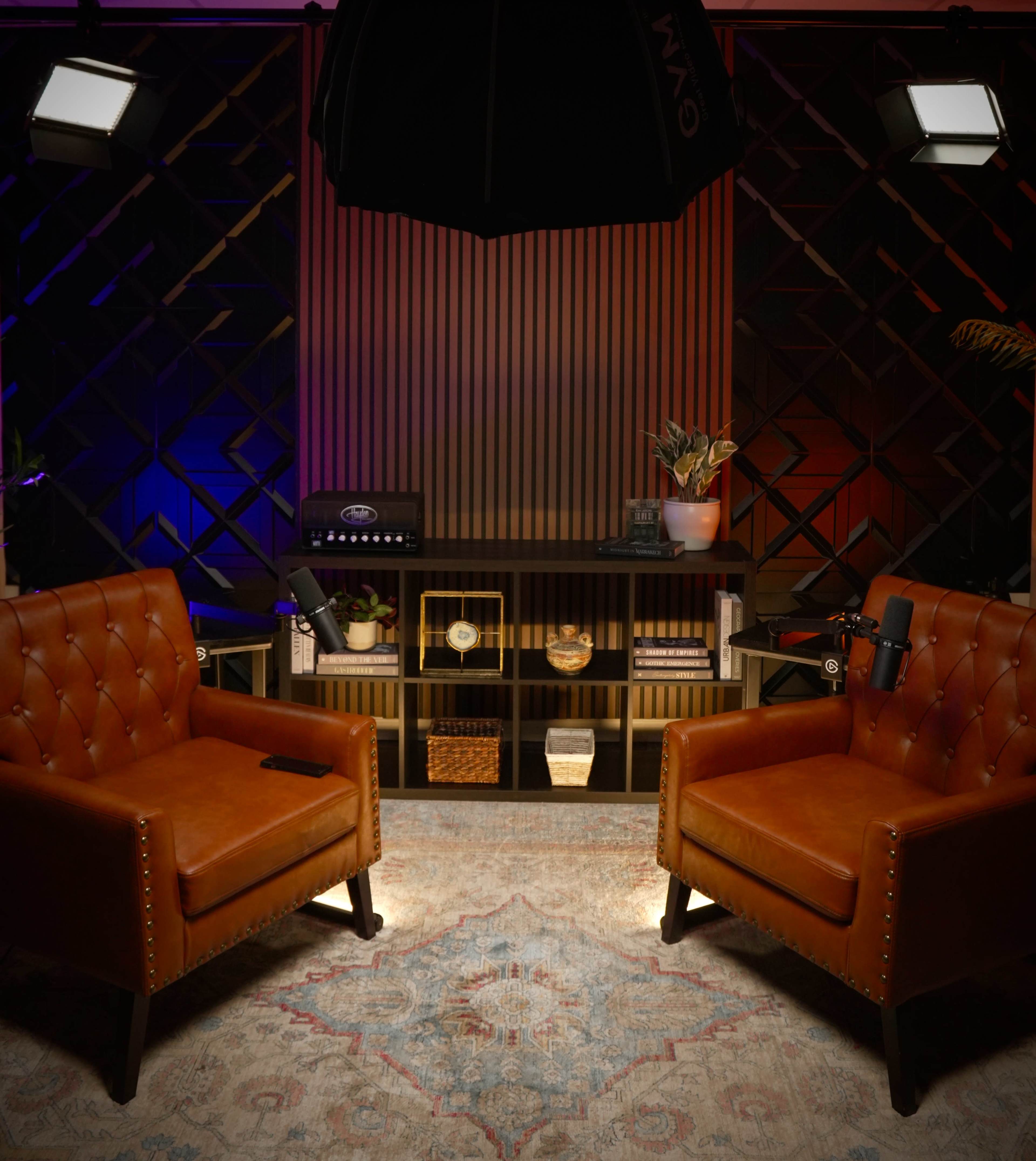 The image shows a cozy studio set with two brown tufted chairs facing a shelf filled with decorative items, framed by dramatic lighting and a textured backdrop.