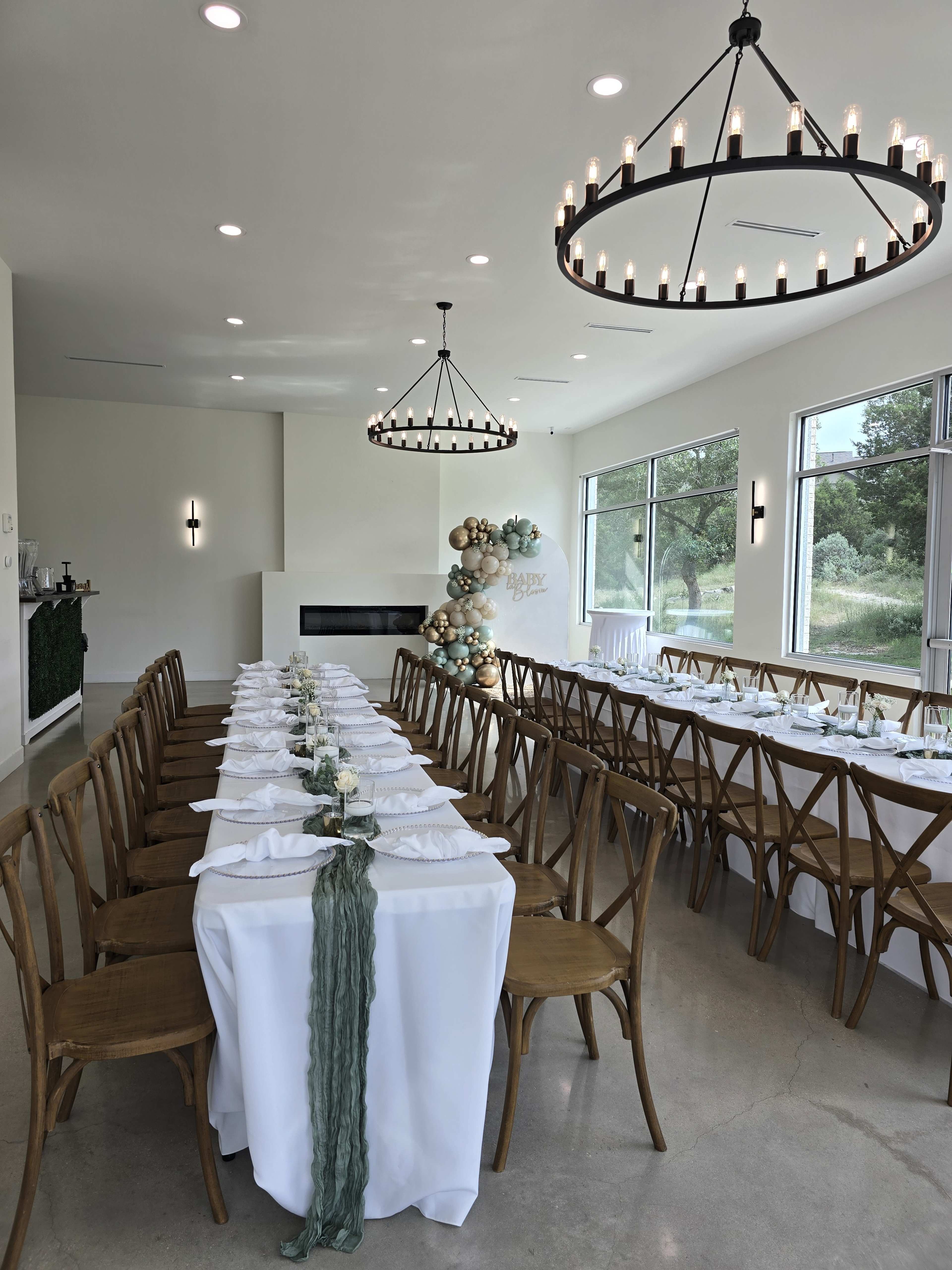 A large, elegantly set dining table with wooden chairs is arranged in a well-lit room featuring two chandeliers and decorative elements.