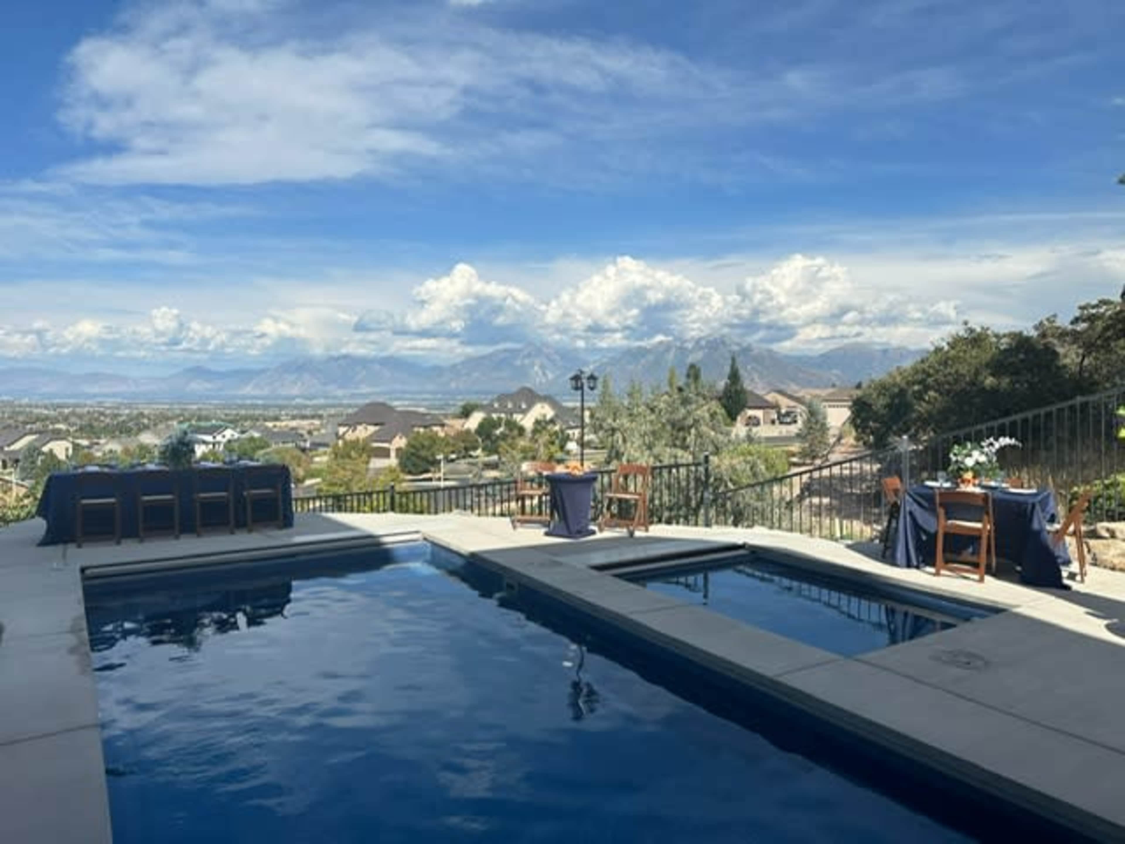 The image shows a swimming pool with adjacent tables set for an outdoor gathering against a backdrop of mountains and a clear sky.