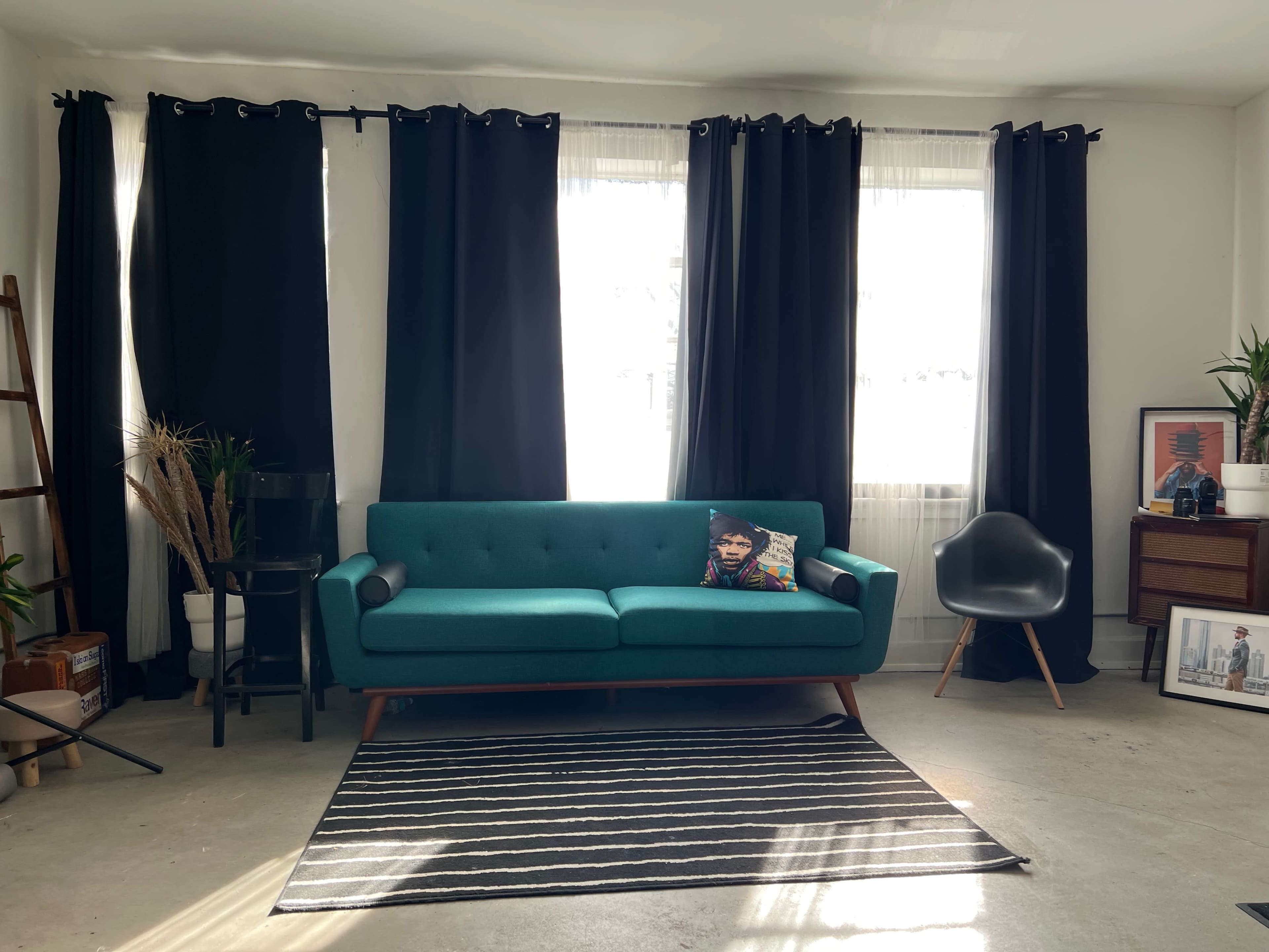 The room features a turquoise sofa with a patterned pillow, black curtains, a striped rug, and various furnishings including a ladder shelf and a chair.