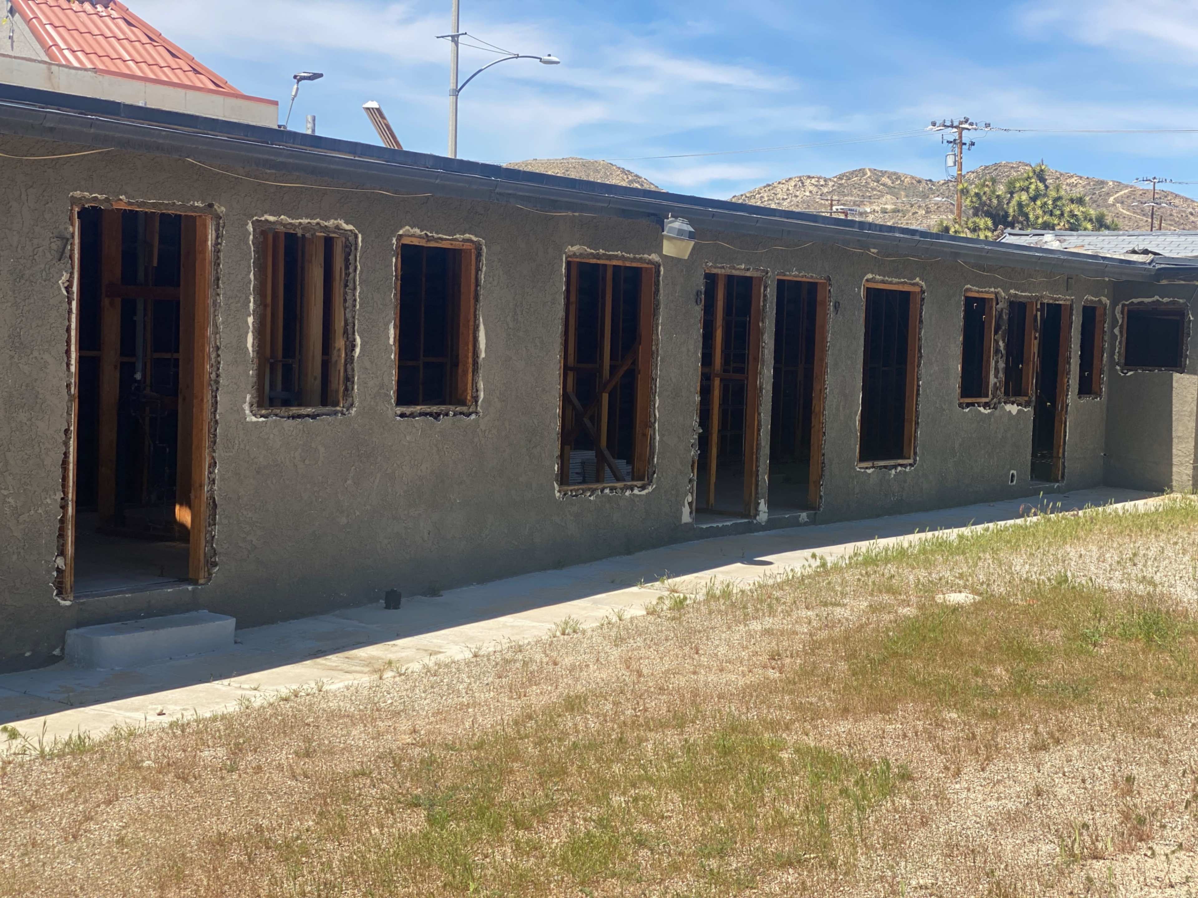 The image depicts an unfinished single-story building with multiple open windows and a dirt pathway alongside a patch of overgrown grass.