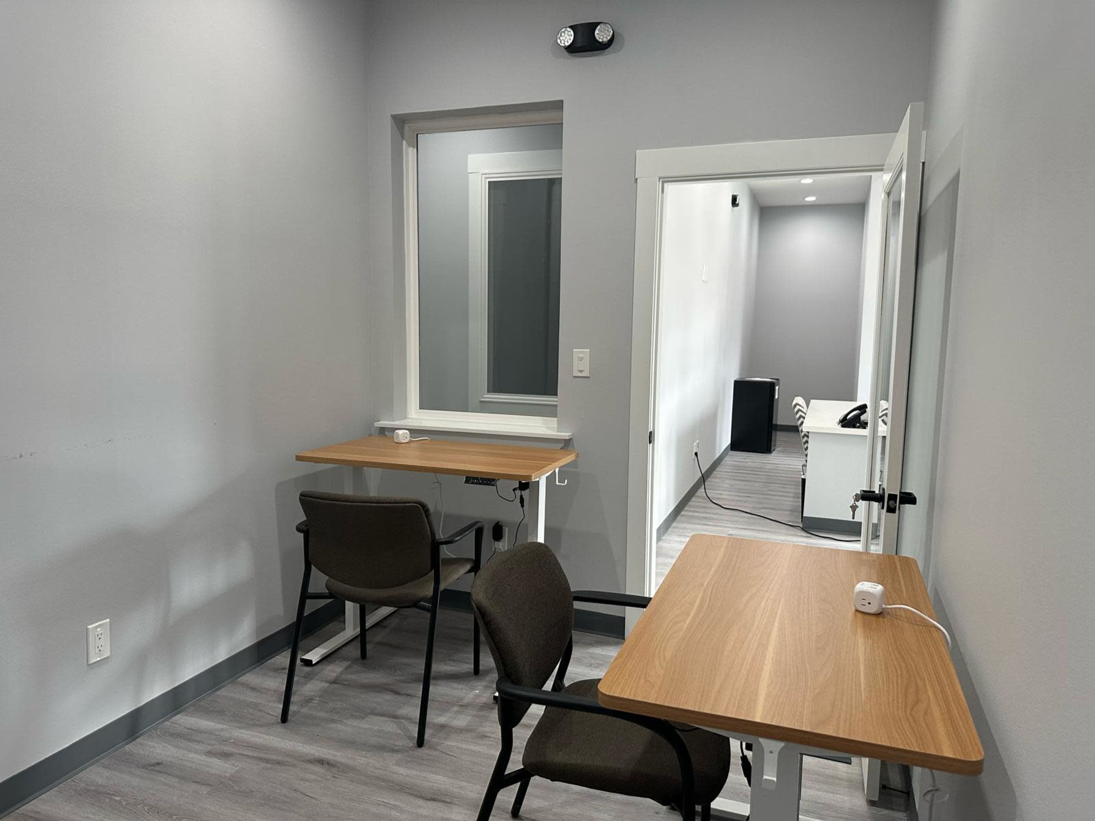 The image shows a small room with two wooden tables and chairs, and an open doorway leading to a light-colored hallway.