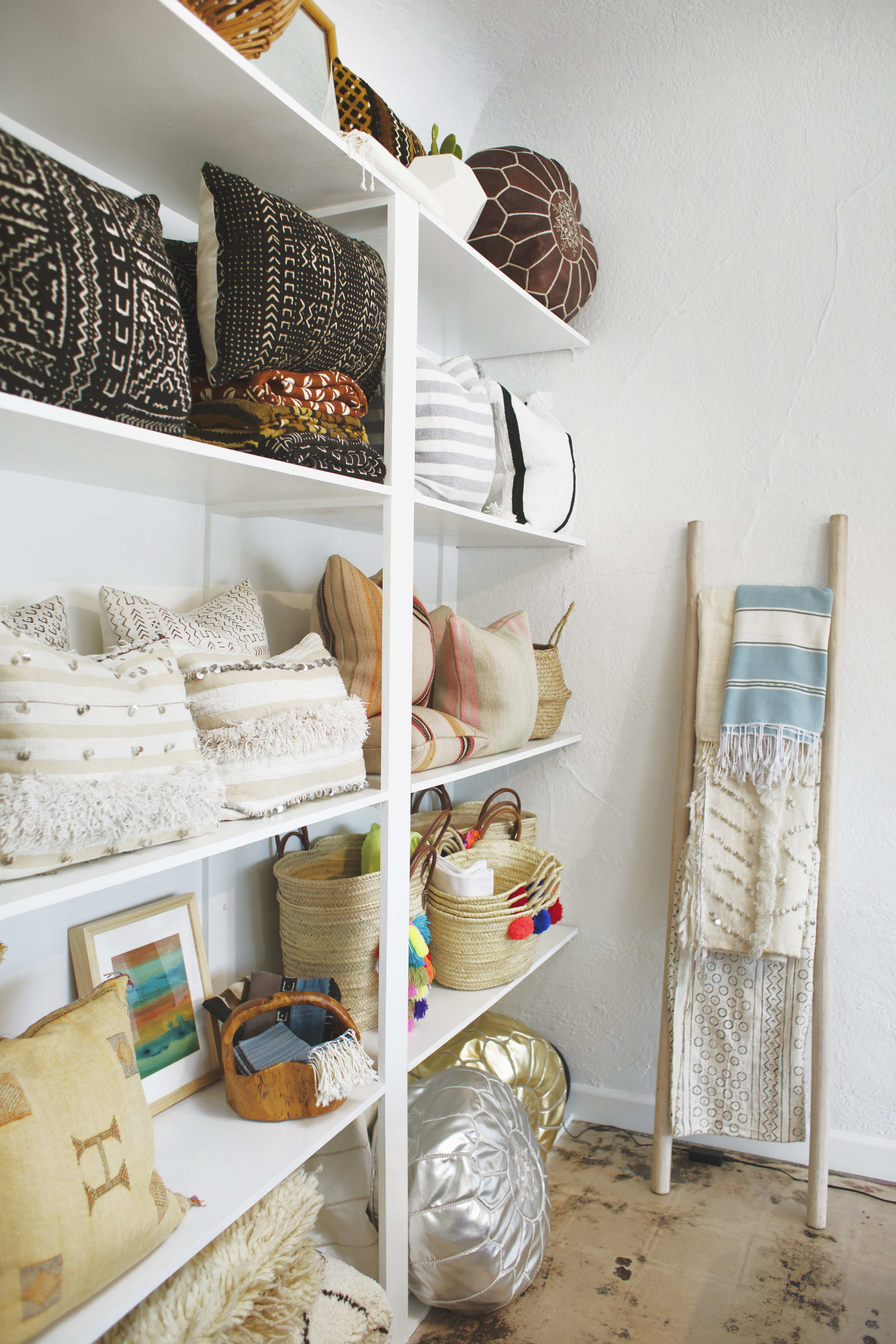A neatly organized display of various decorative pillows and woven baskets on white shelves, alongside a textile wall hanging.