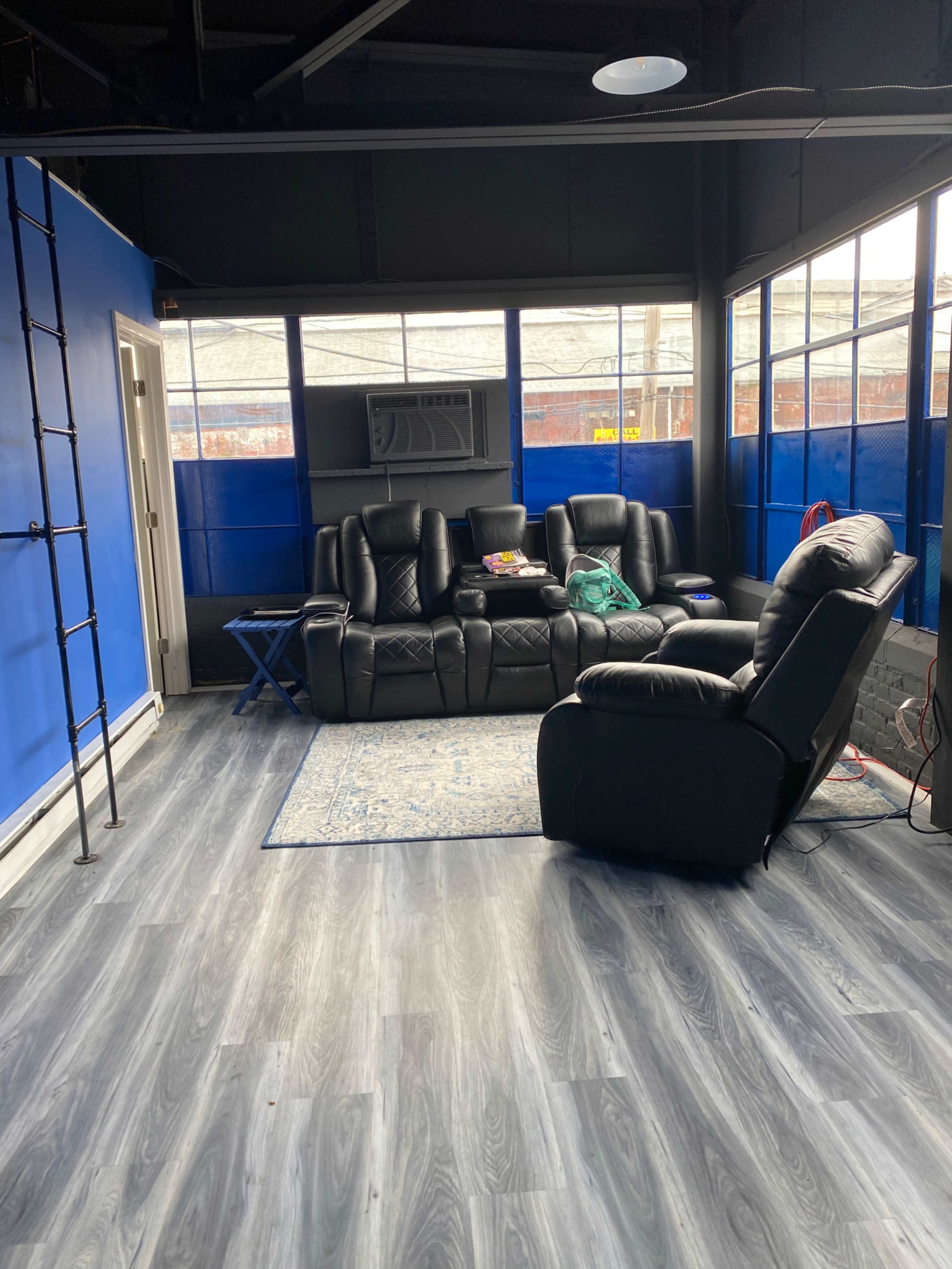 The image shows a room with blue walls, black leather seating arranged around a small table, and large windows letting in natural light.