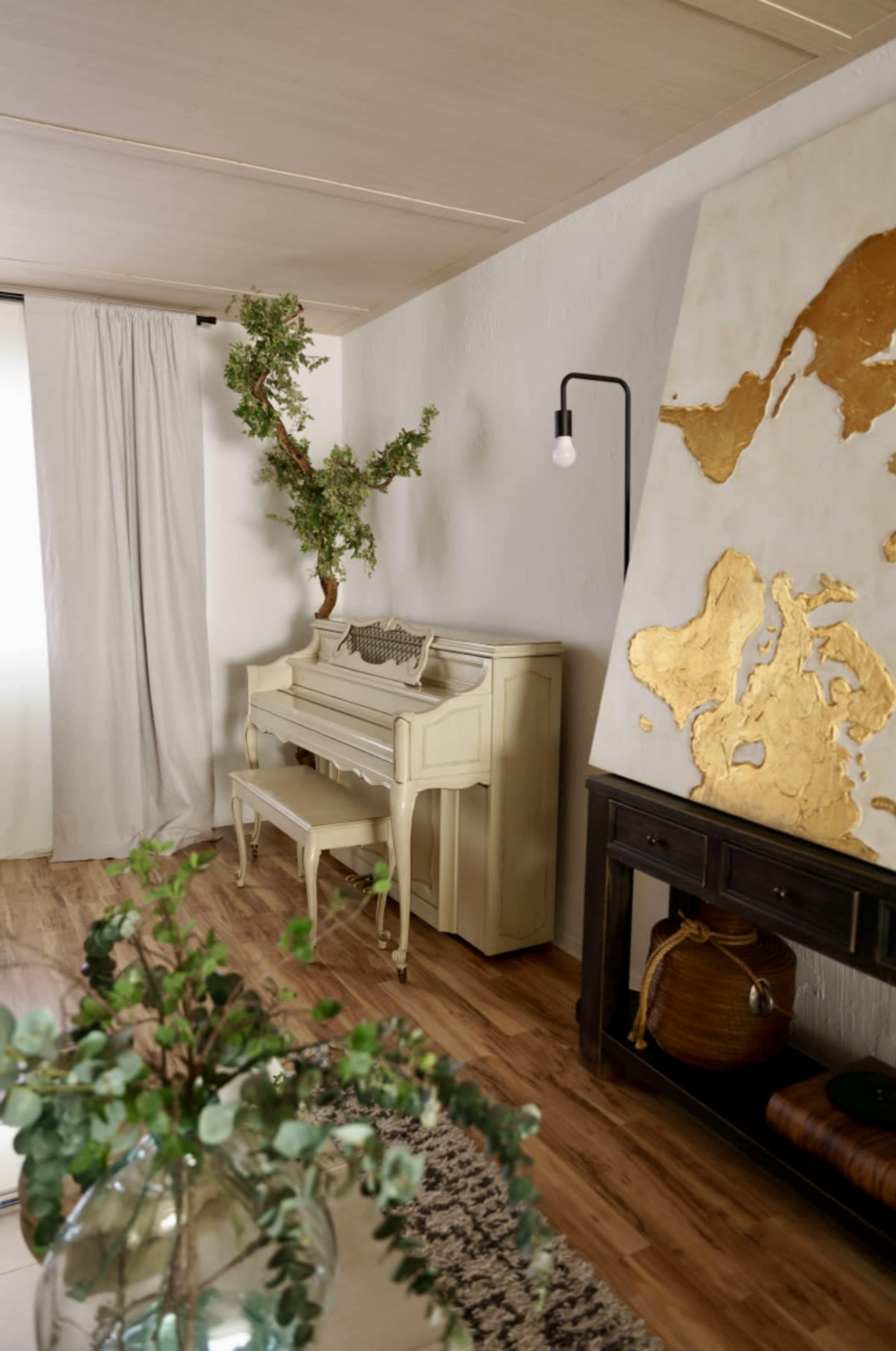 A vintage piano is situated beside a wall featuring a large, gold-leaf world map, with a decorative plant placed above it in a neutral-colored room.