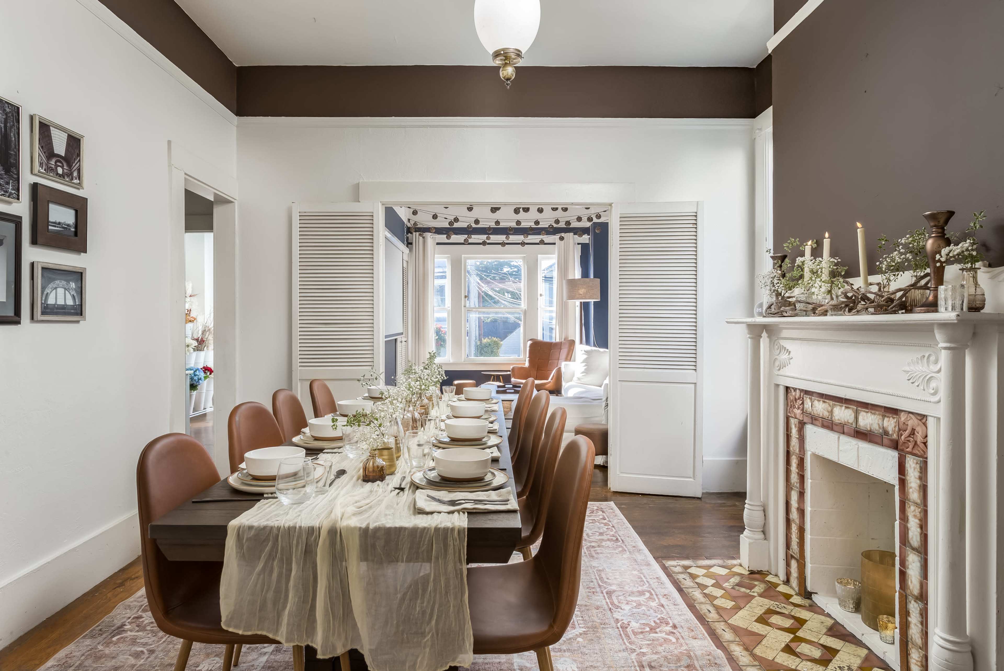 A dining room features a long table set for a meal with eight brown leather chairs, surrounded by white walls and a dark brown accent wall, and a decorative fireplace in the background.