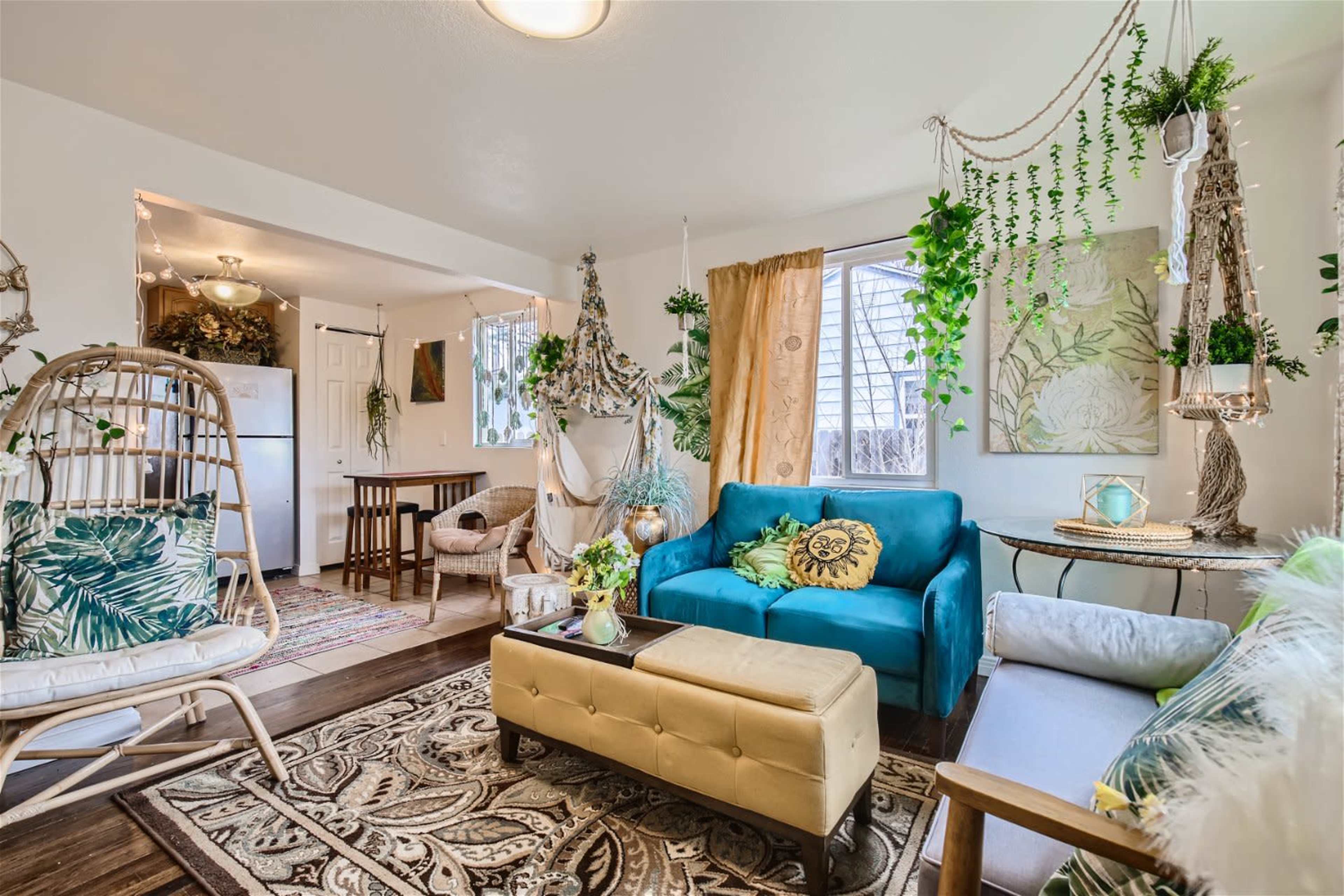 The living room features a blue couch, a yellow ottoman, and various plants hanging from the ceiling and walls.