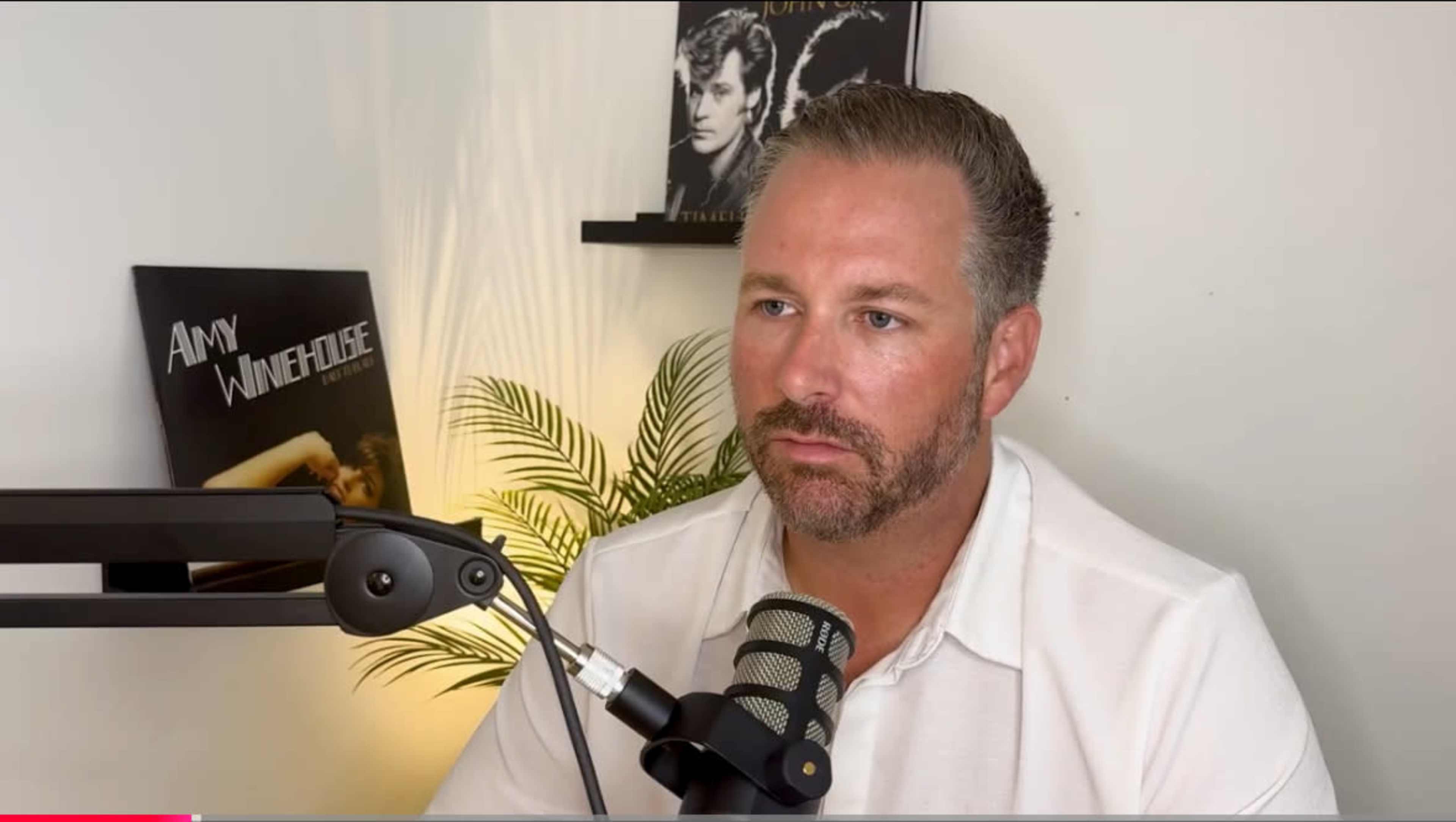 A man sits in front of a microphone, with album covers of Amy Winehouse and a male artist visible on the wall behind him.
