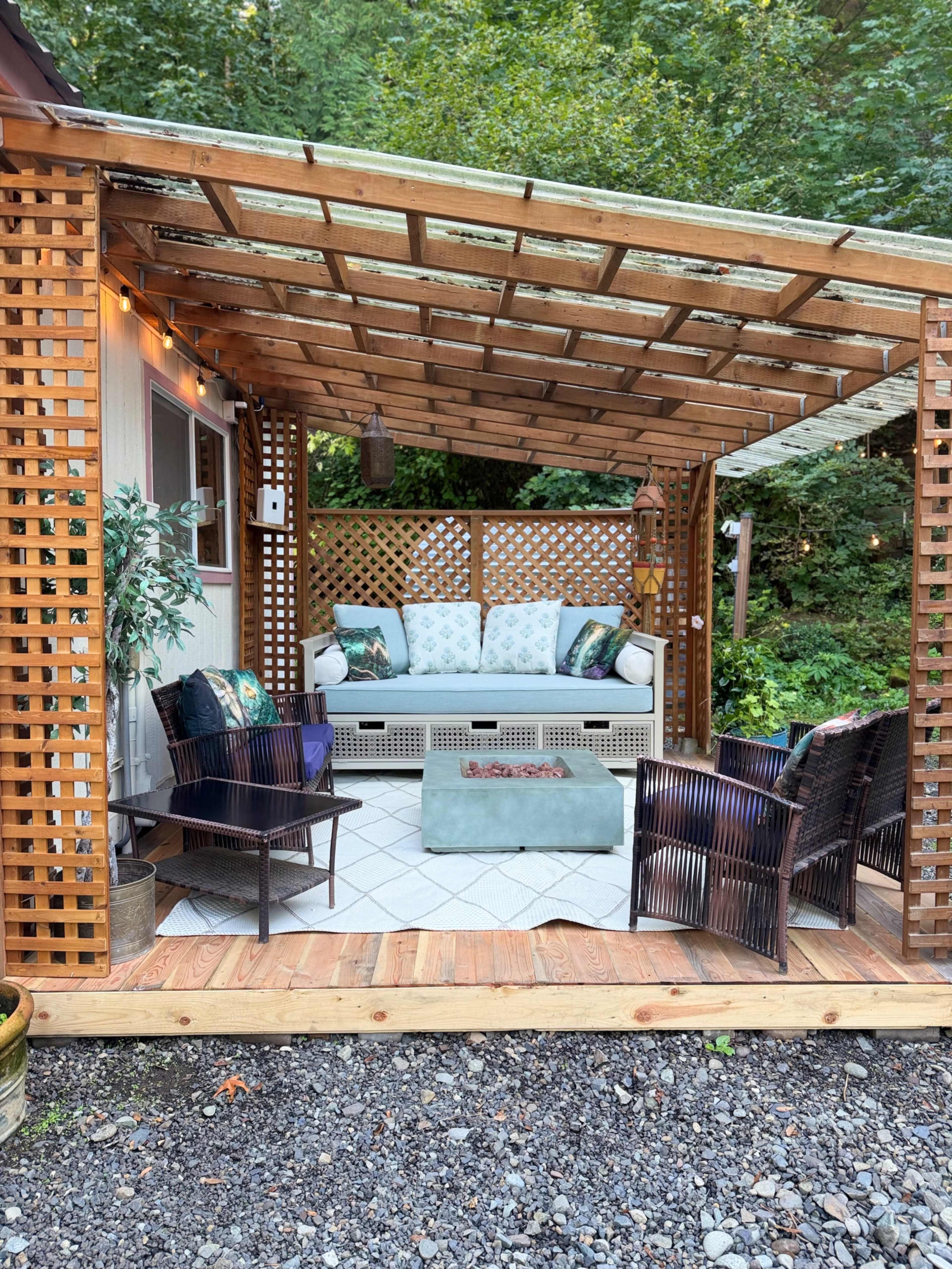 A cozy outdoor seating area features a wooden pergola with comfortable furniture, including a sofa and chairs, surrounded by greenery.
