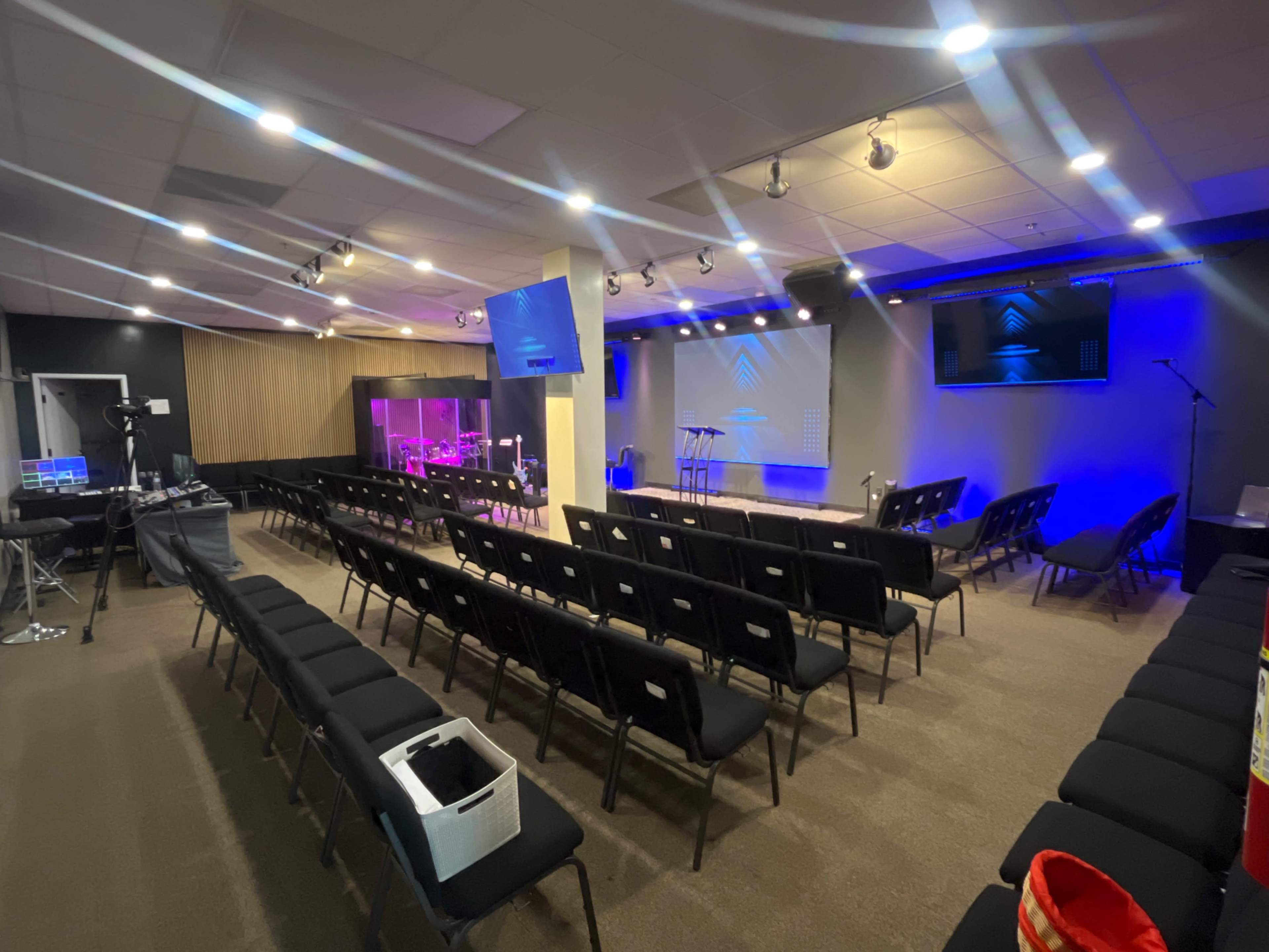 The image shows a modern church or auditorium space with rows of black chairs facing a stage that features lighting and a projection screen.
