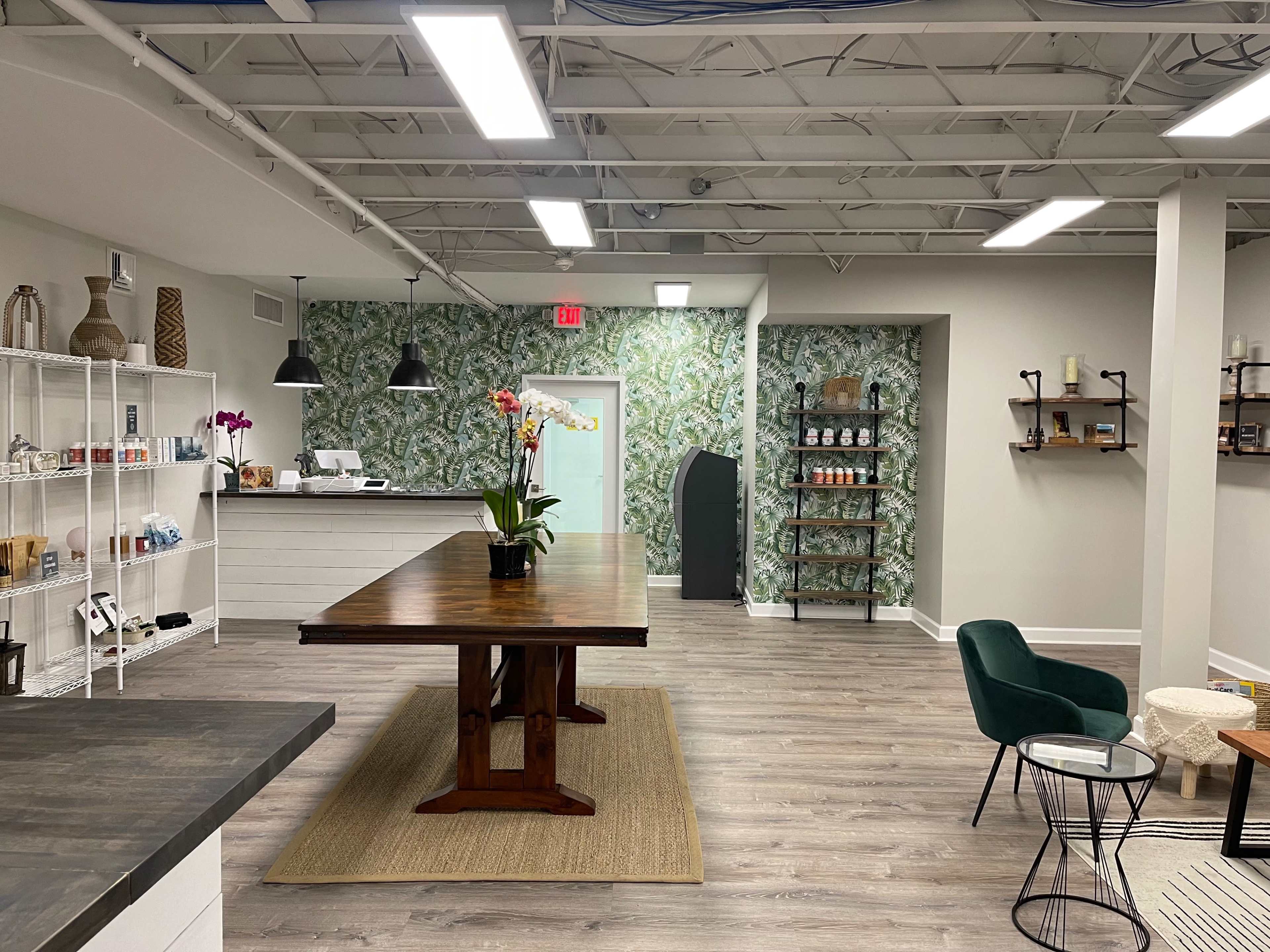 A modern interior space with a large wooden table, shelves filled with various products, a green accent wall, and minimalist furniture.
