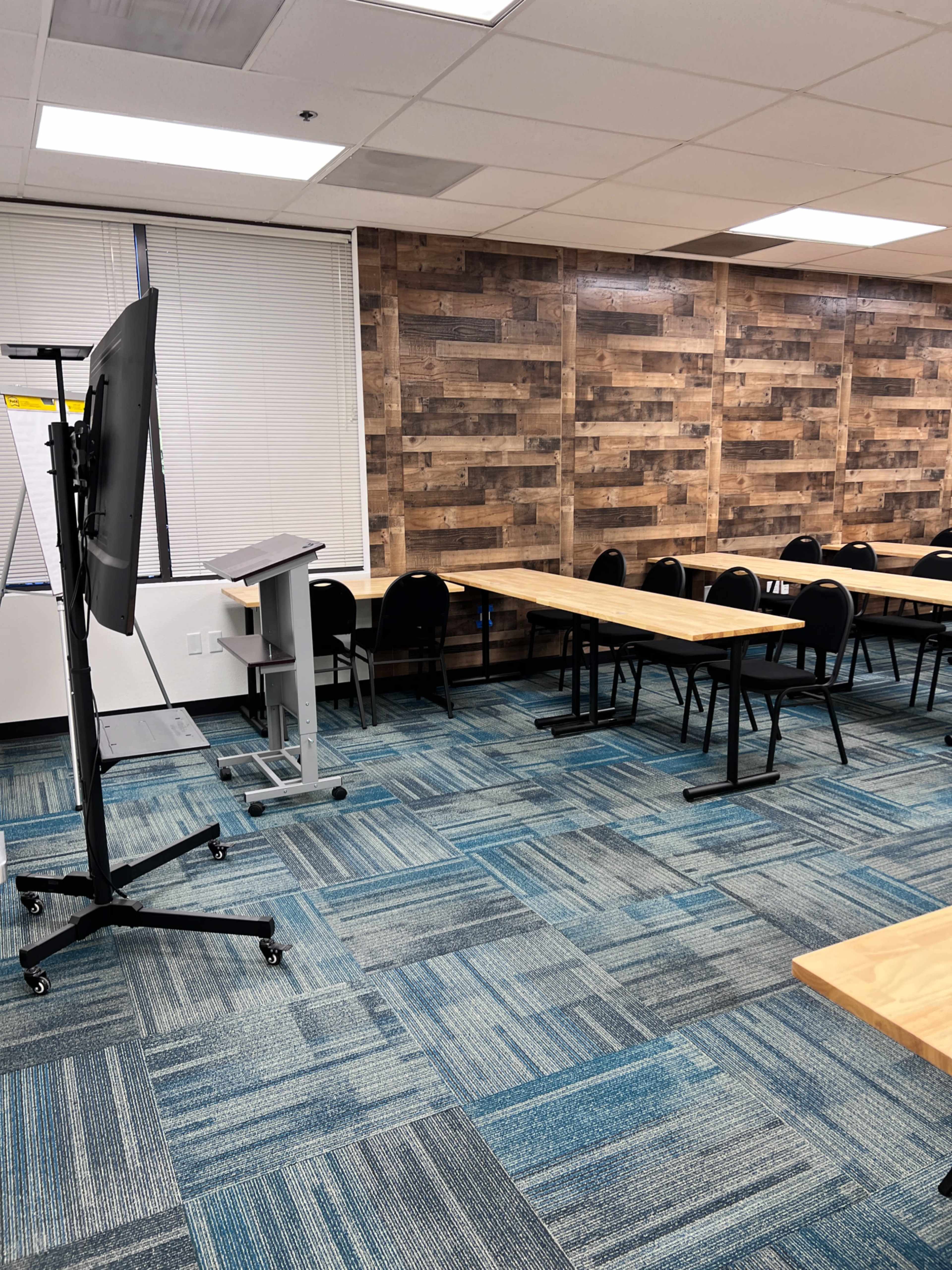 A classroom with wooden tables arranged in rows, black chairs, a presentation screen, and a podium against a paneled wall.