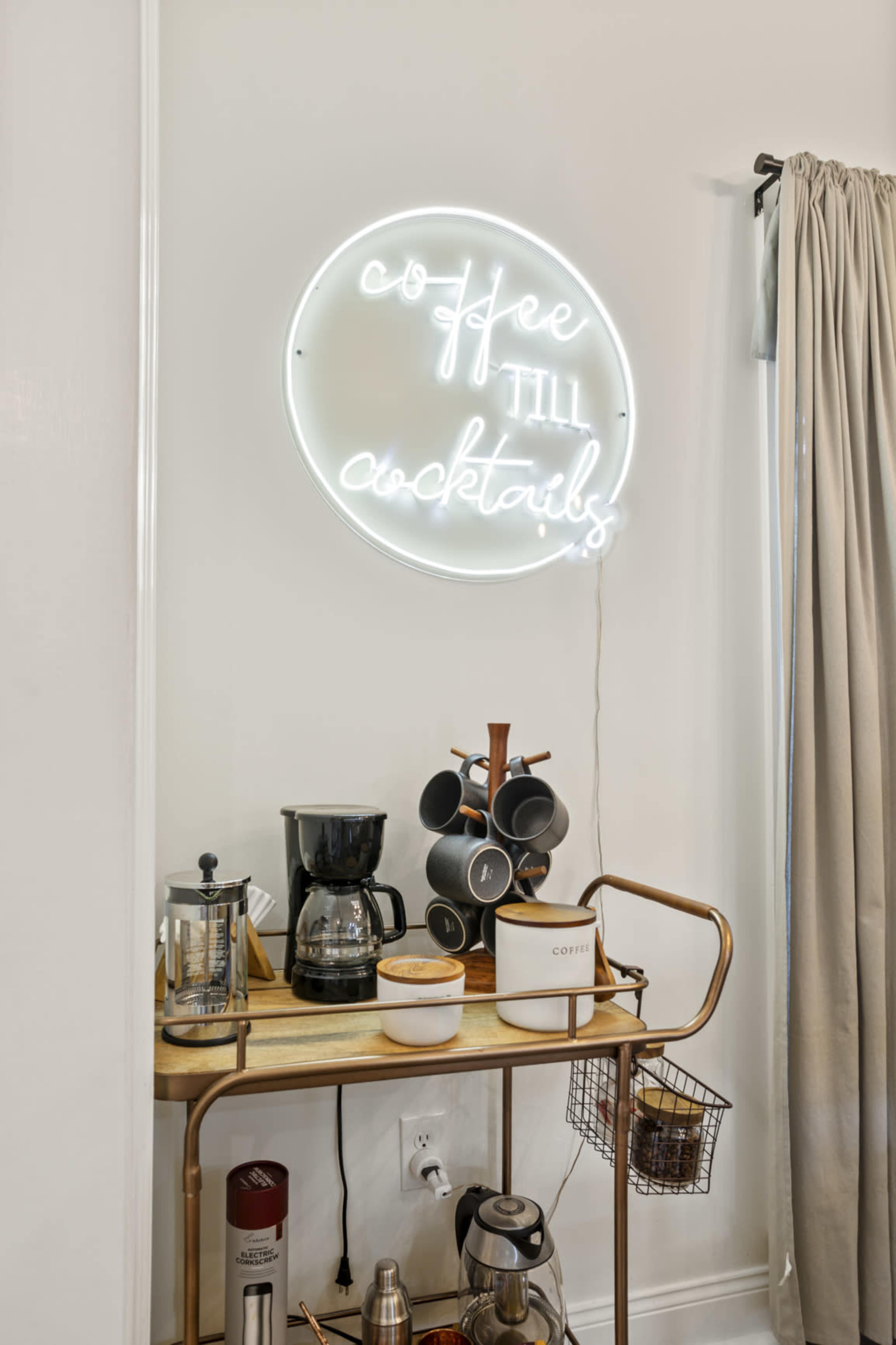 A neon sign that reads "coffee till cocktails" hangs above a wooden cart with a coffee maker, a French press, and several cups.
