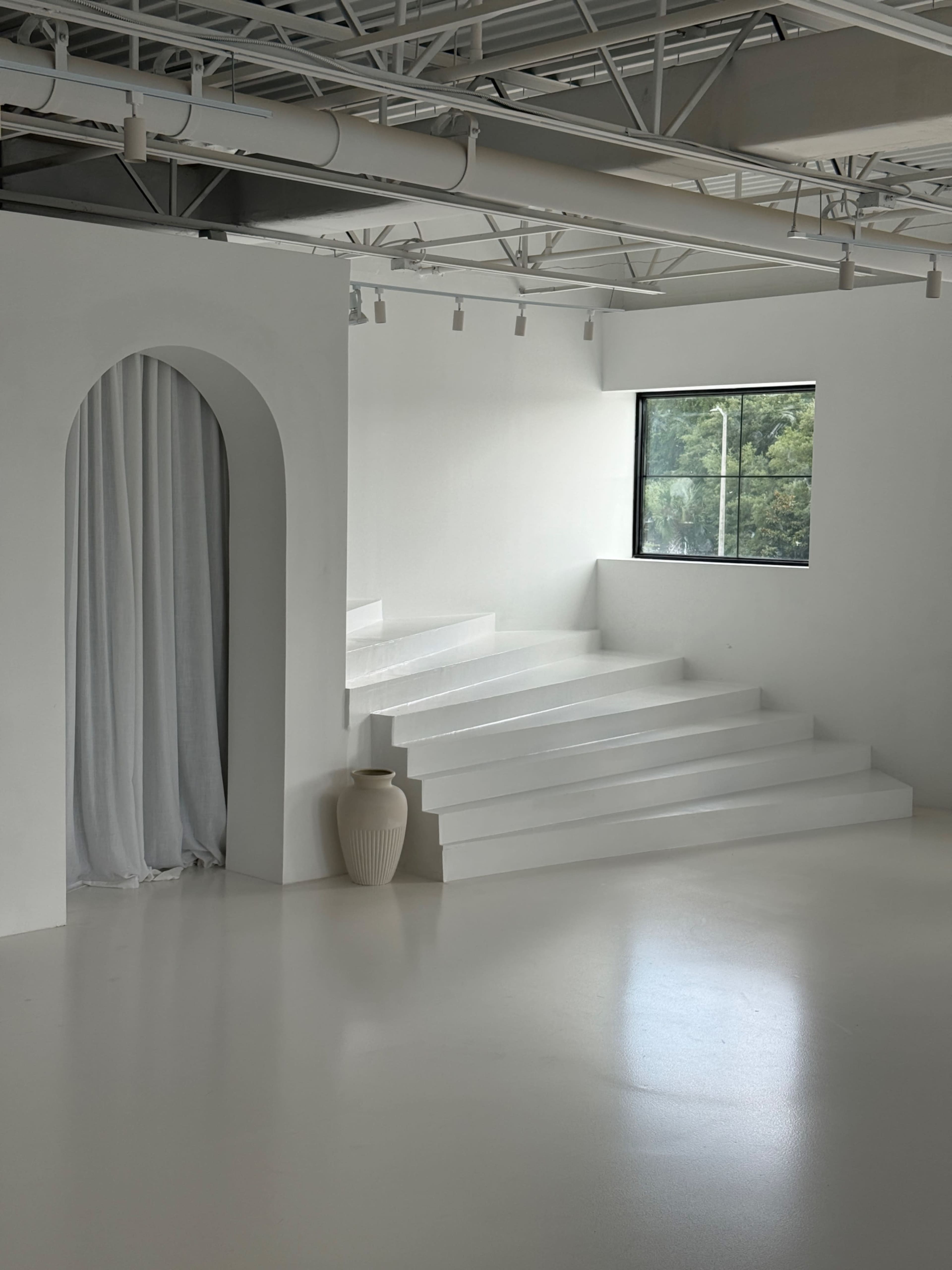 The image shows a minimalist interior space featuring steps leading up to a large window, an archway with curtains, and a decorative vase.