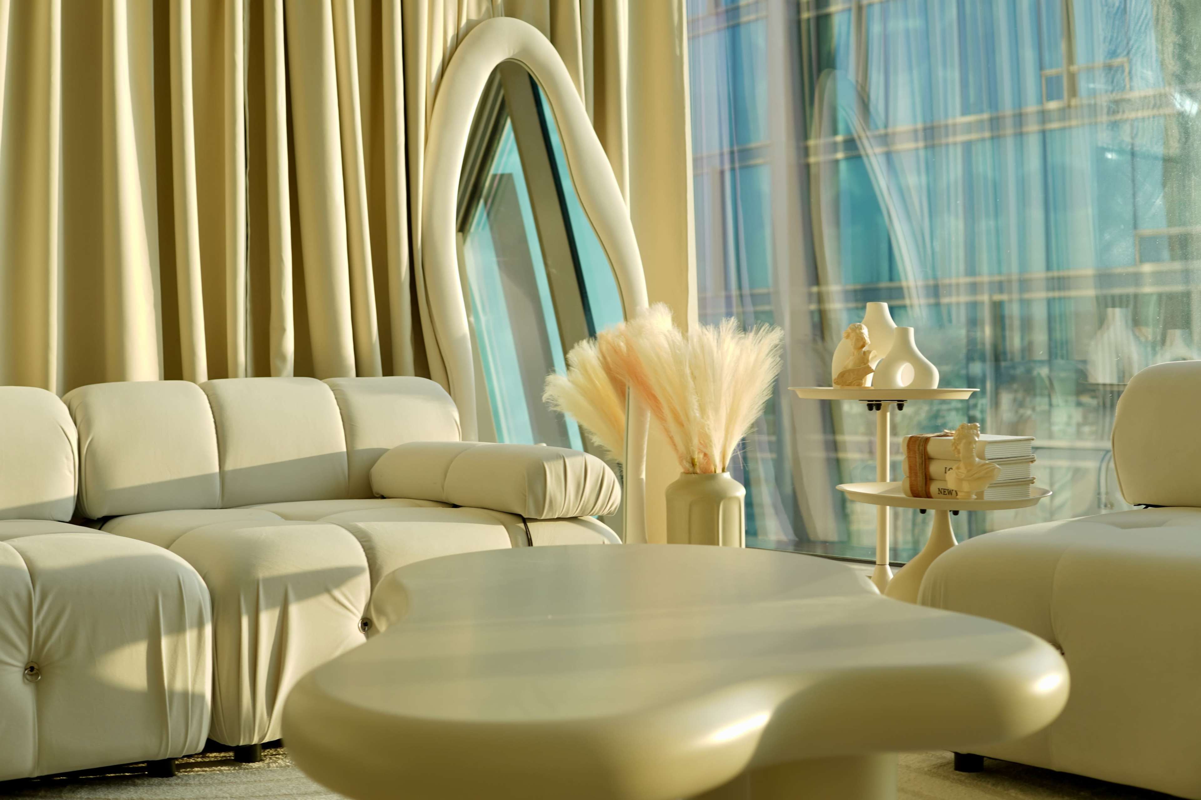 A modern interior with a light-colored sofa, a sculptural coffee table, and a decorative vase with dried flowers, all set against large windows with sheer curtains.