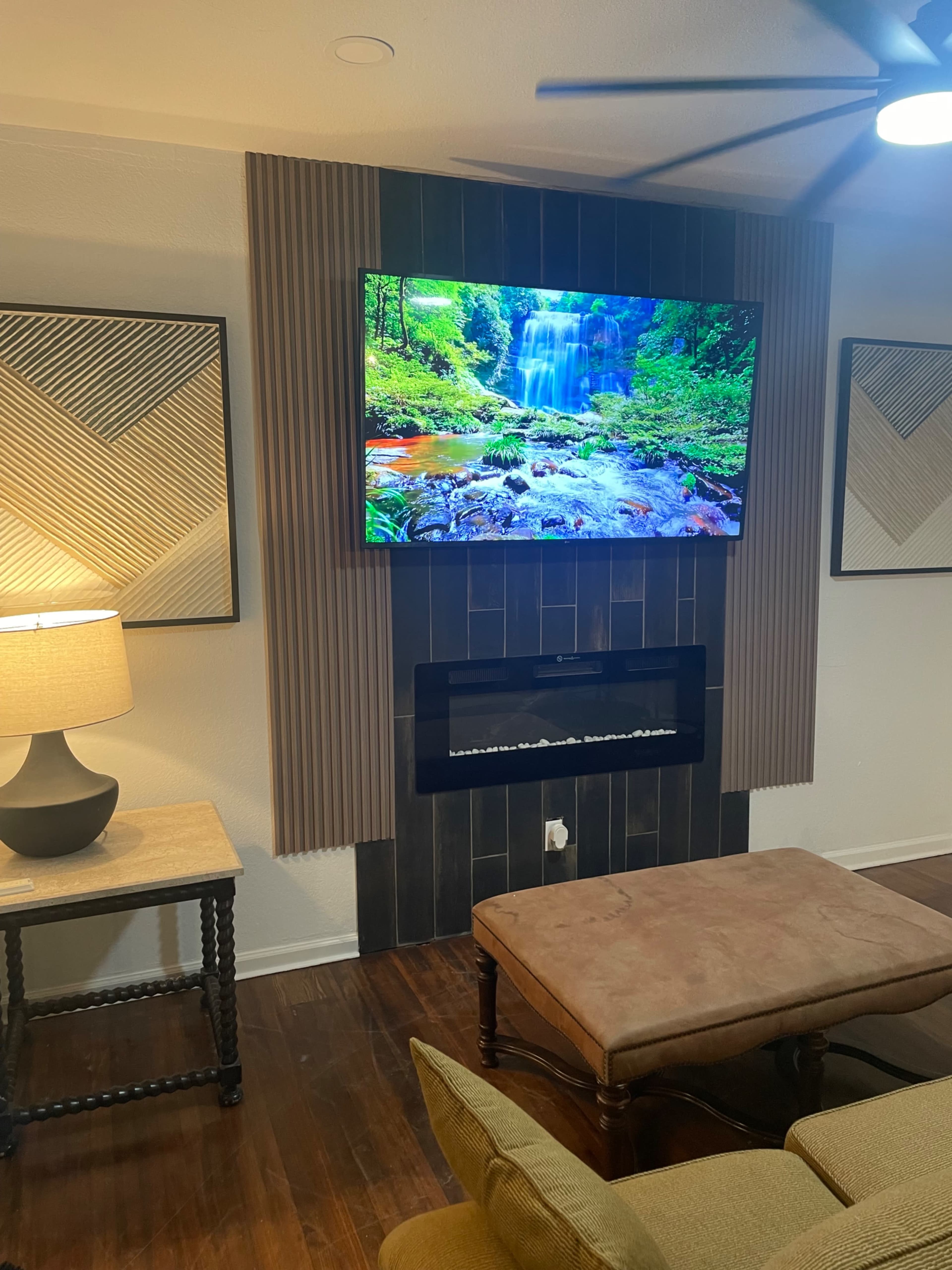 A television displaying a waterfall scene is mounted on a wooden feature wall above a modern fireplace, with a lamp and a coffee table in the foreground.