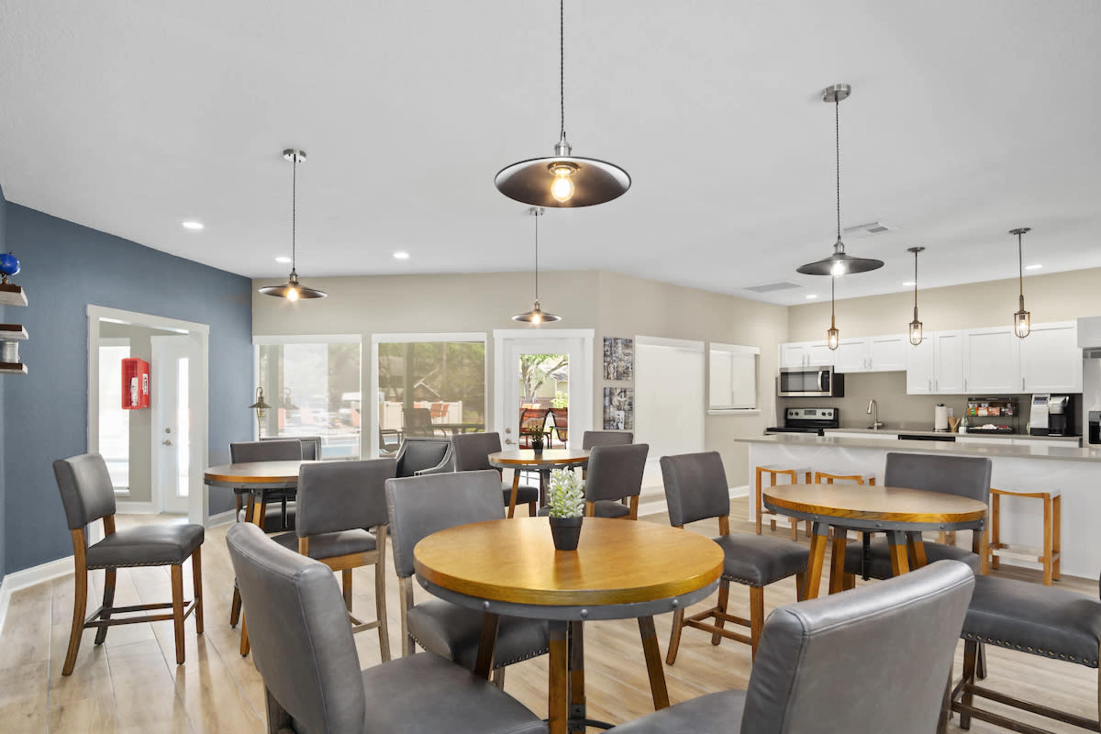 A modern communal space features round wooden tables surrounded by gray upholstered chairs, with overhead pendant lights and an open kitchen area in the background.