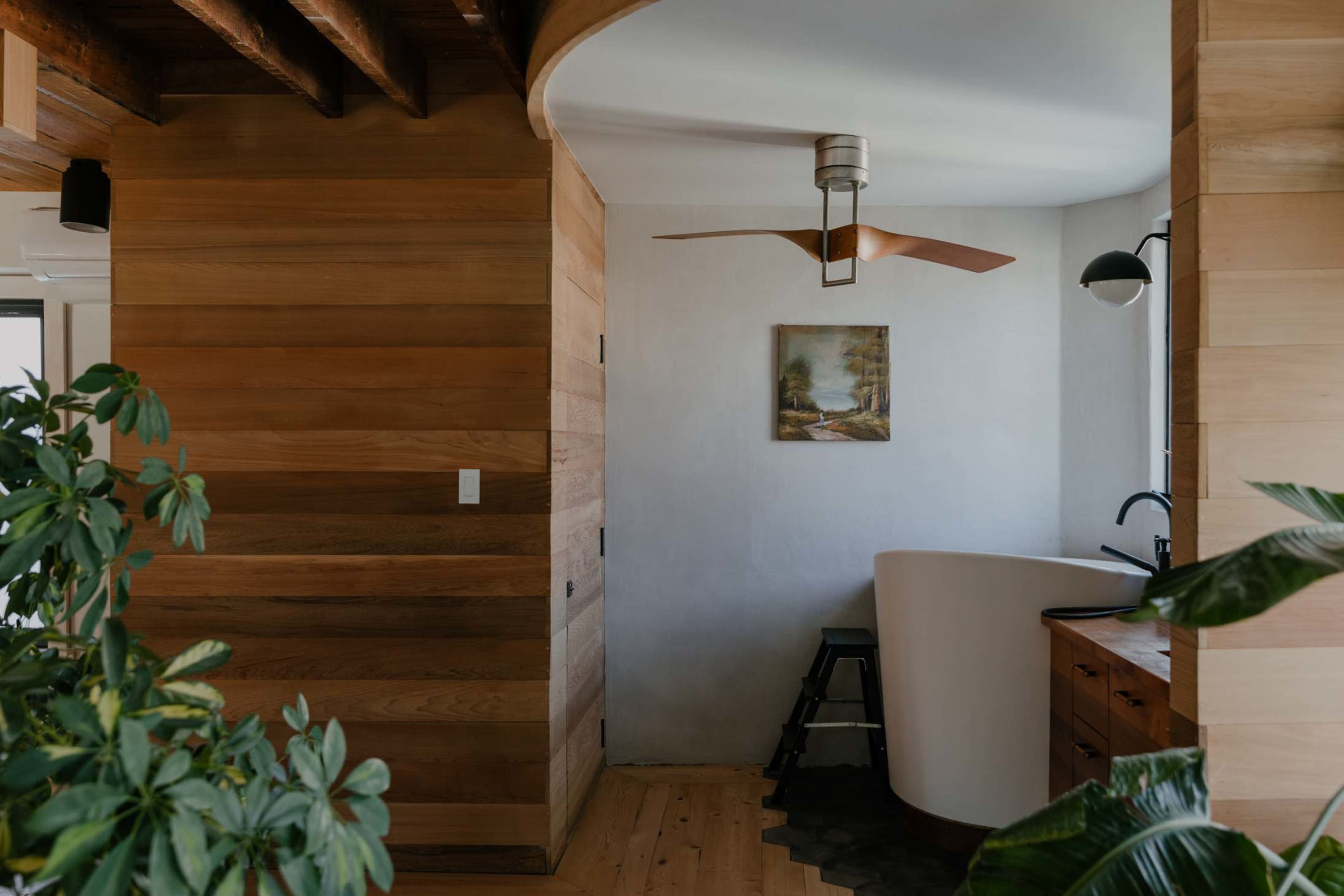 A cozy interior space featuring wooden walls, a ceiling fan, a small painting, and a curved wall leading to a kitchen area.