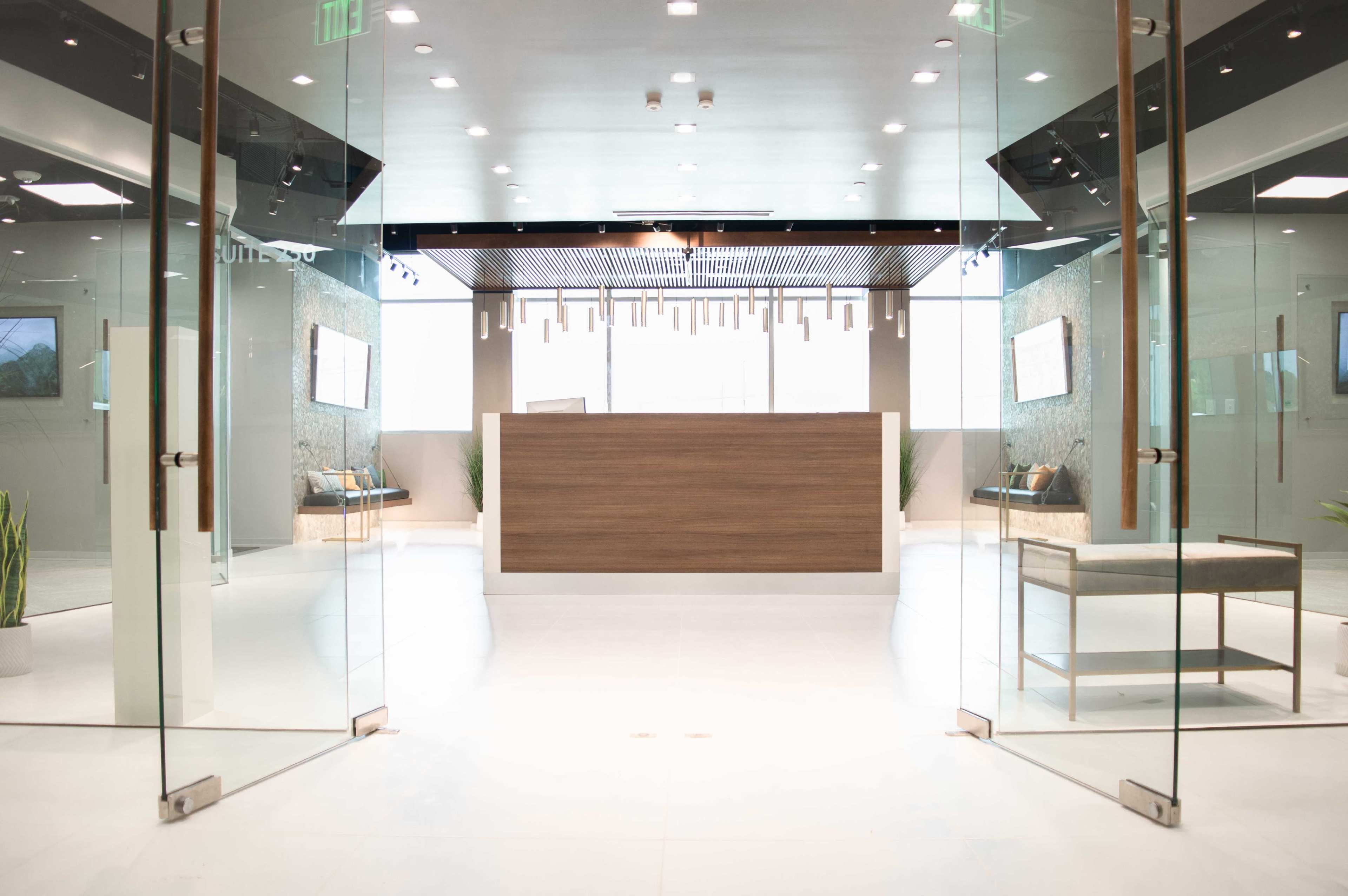 The image shows a modern reception area with a wooden front desk, glass doors, and minimalist decor.