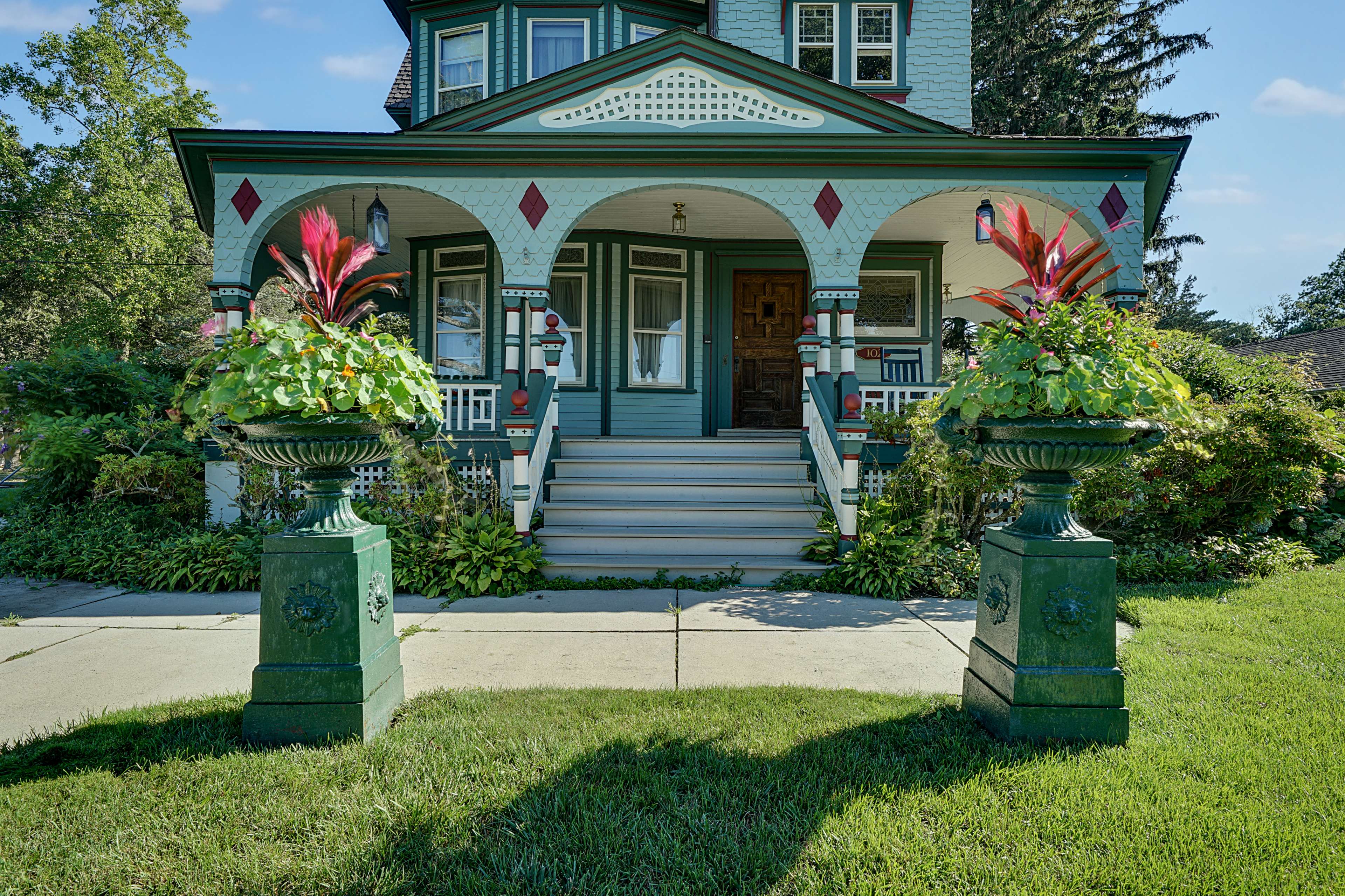 A two-story house with a turquoise exterior, adorned with decorative trim and flanked by large planters filled with vibrant foliage on either side of a front staircase.