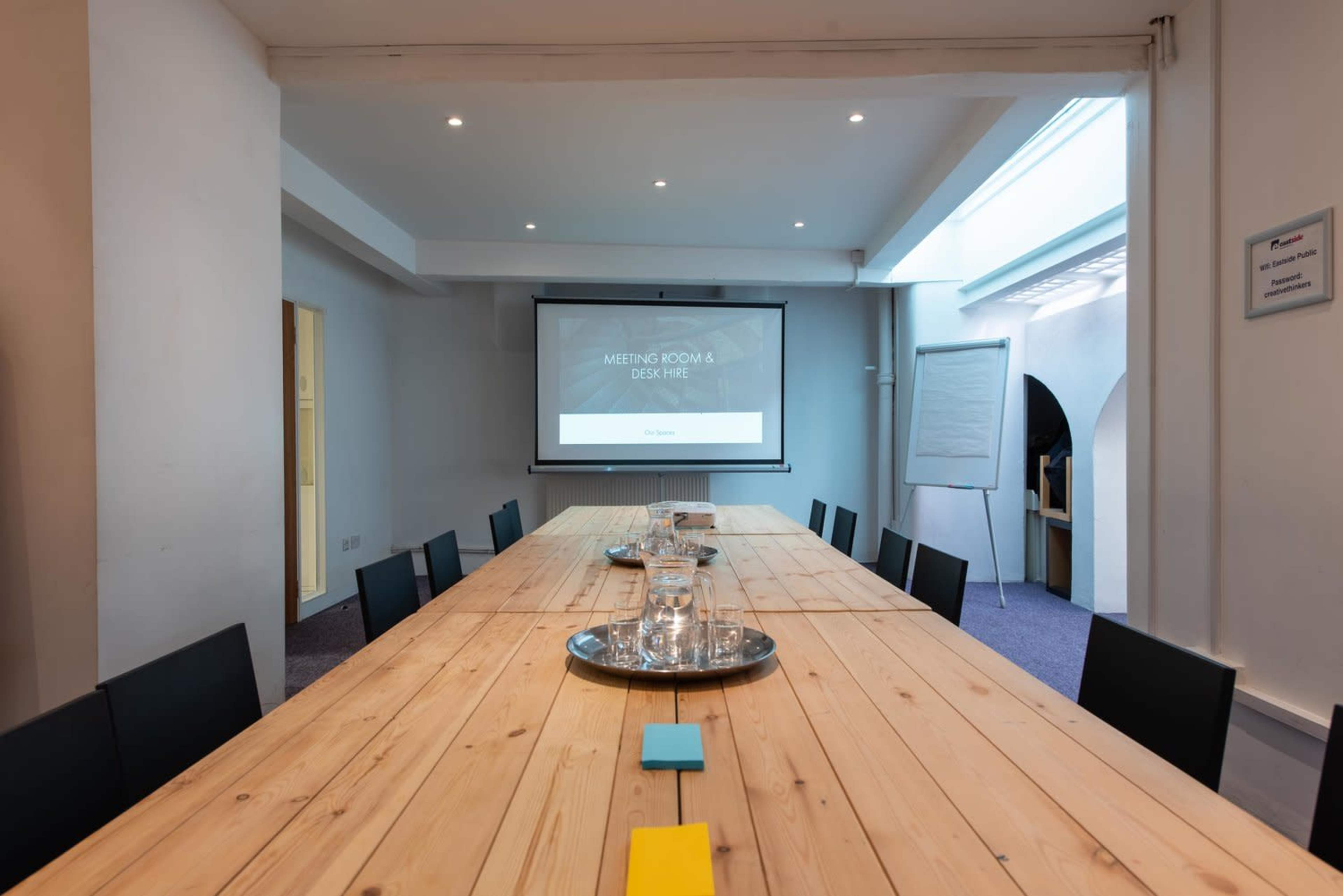 A spacious meeting room features a long wooden table, a projector screen displaying "MEETING ROOM & DESK HIRE," and a flip chart.