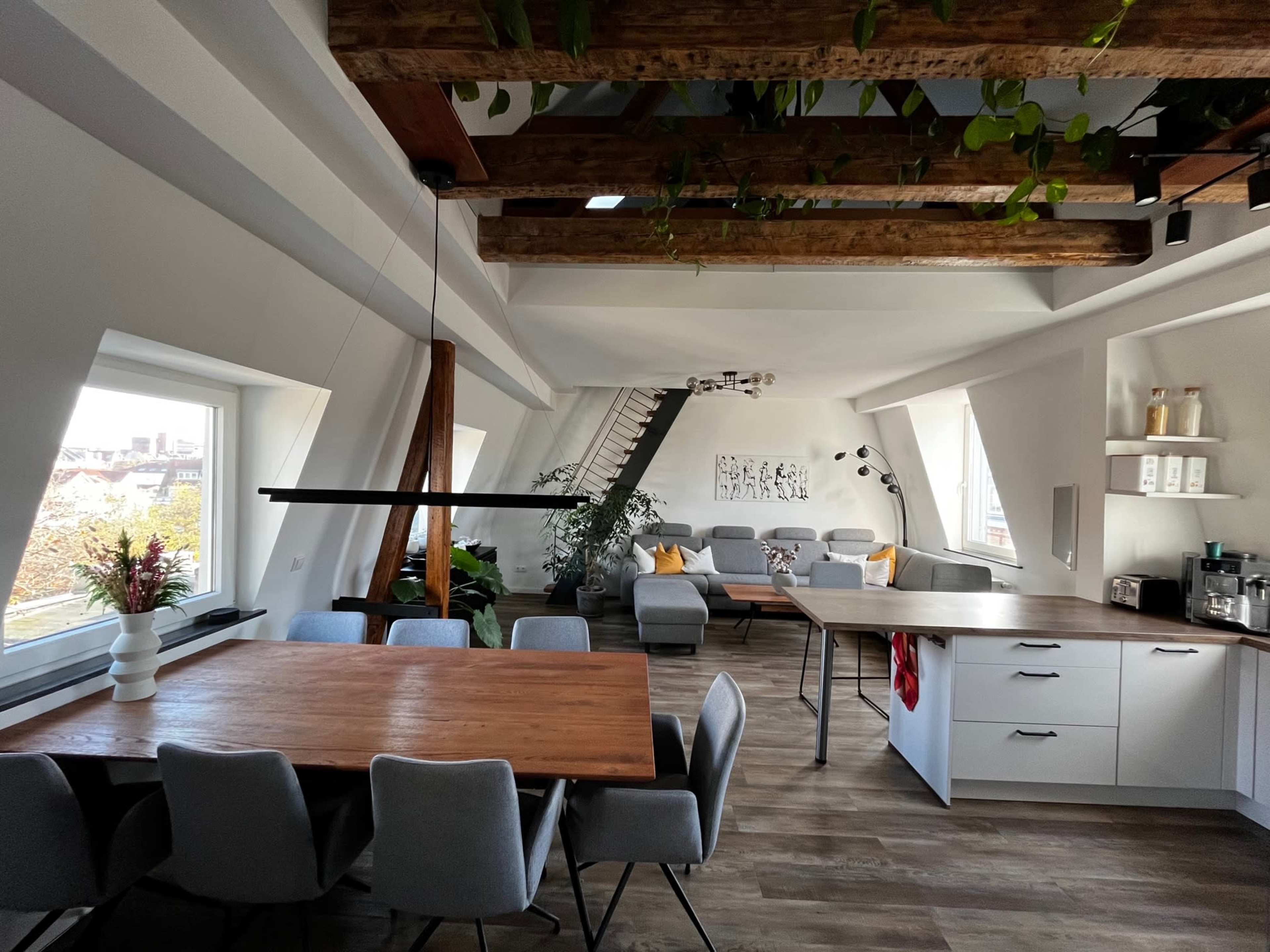An open-concept living space featuring a dining area with a large table, a kitchen with white cabinetry, and a cozy seating area adorned with plants and large windows.