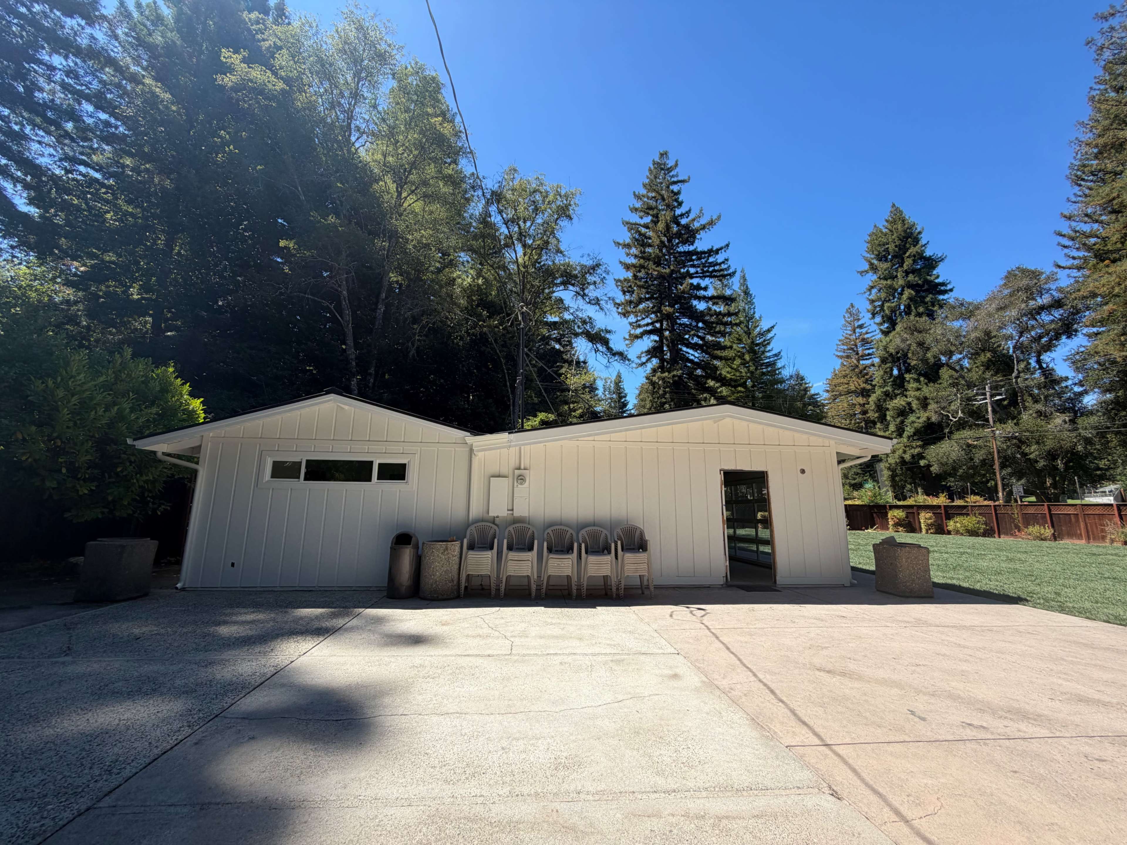 A white, single-story building with a concrete driveway is surrounded by tall trees and features a large window on one side.
