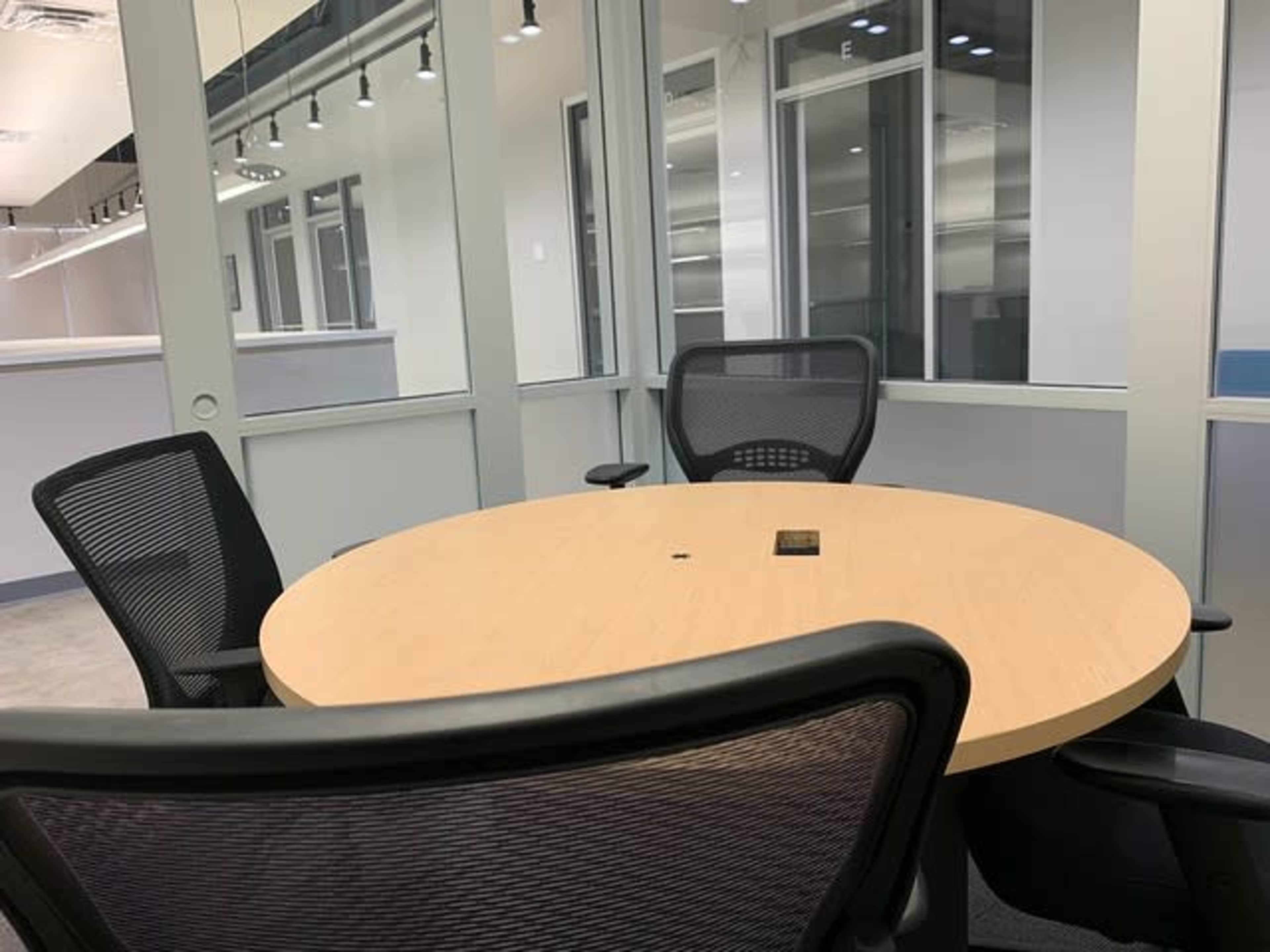 A round wooden table with three black office chairs is set in a meeting room featuring glass walls.