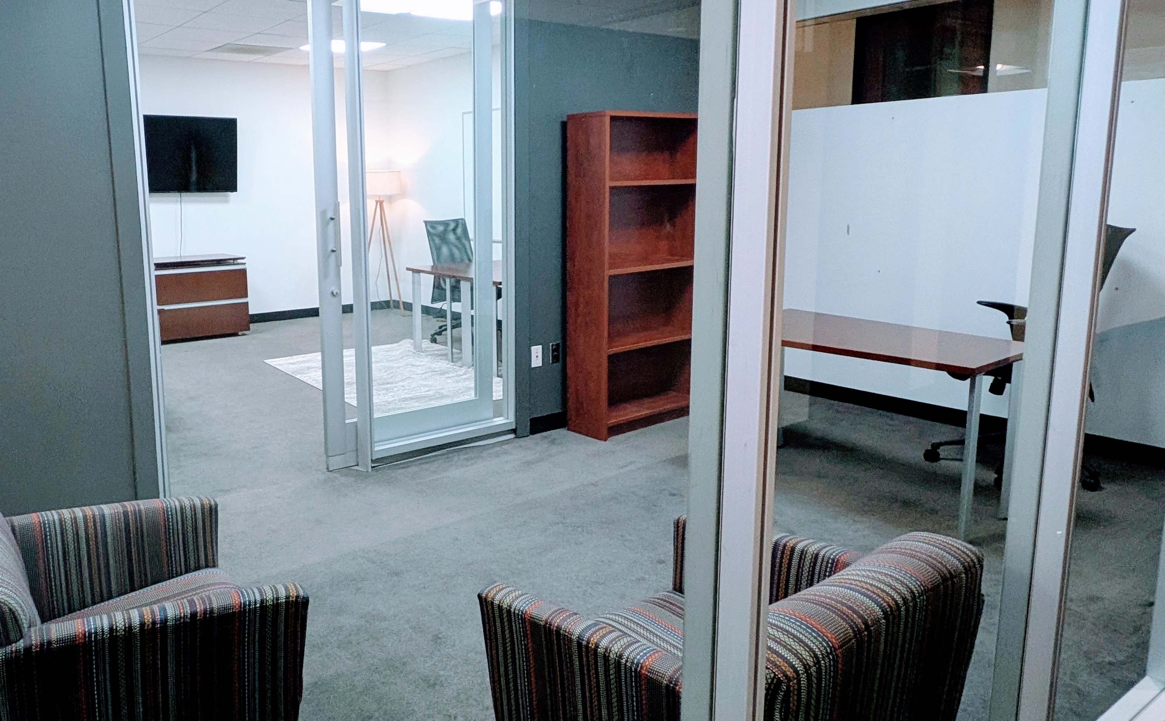 The image shows a modern office setup featuring a glass-walled meeting space, a desk with a chair, a bookshelf, and a seating area with striped armchairs.