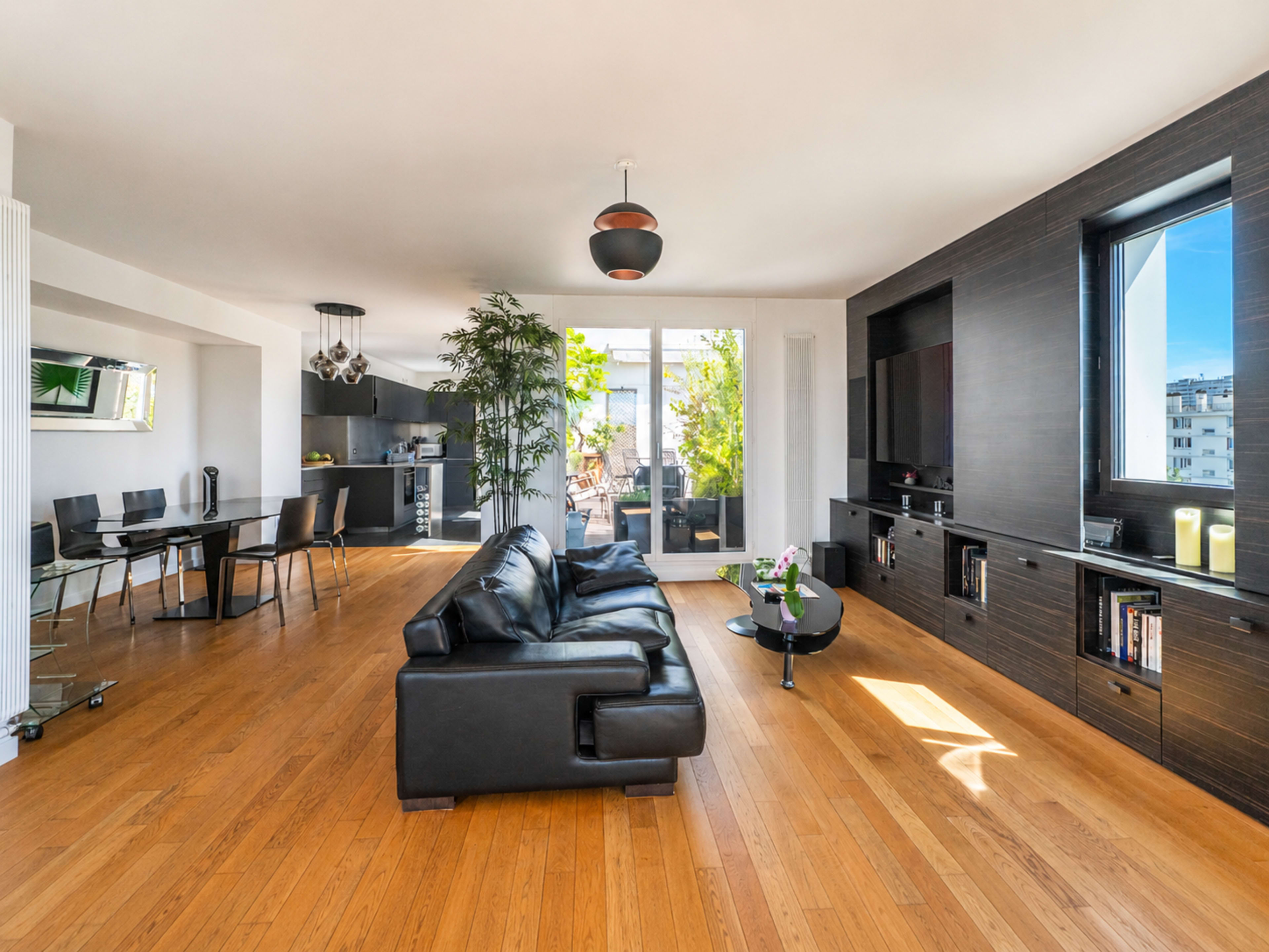 The image shows a modern living room with a black sofa, wooden flooring, and large windows that provide natural light.