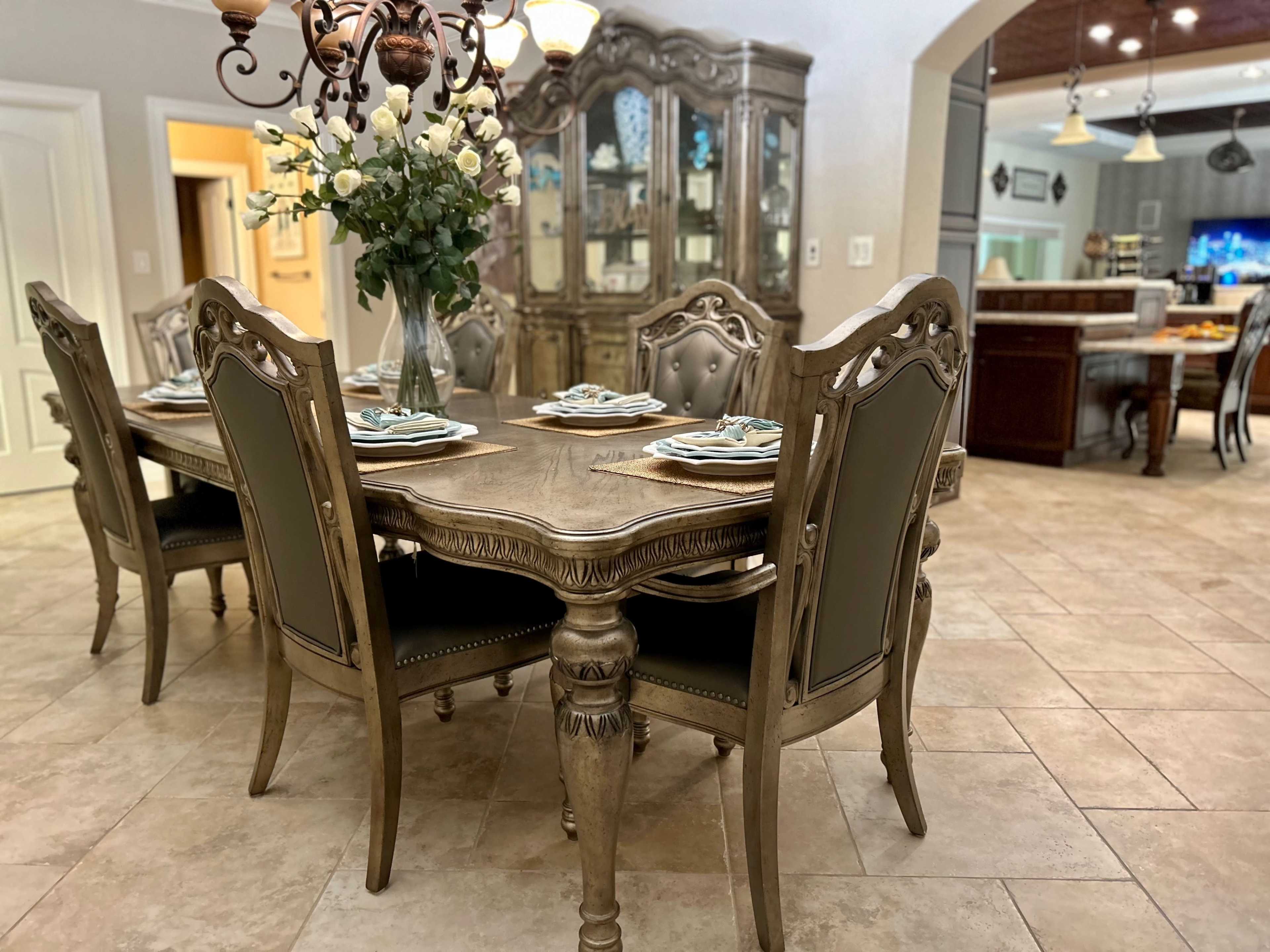 A dining room features a large wooden table set with plates and a floral centerpiece, surrounded by ornate chairs, with a cabinet and kitchen in the background.