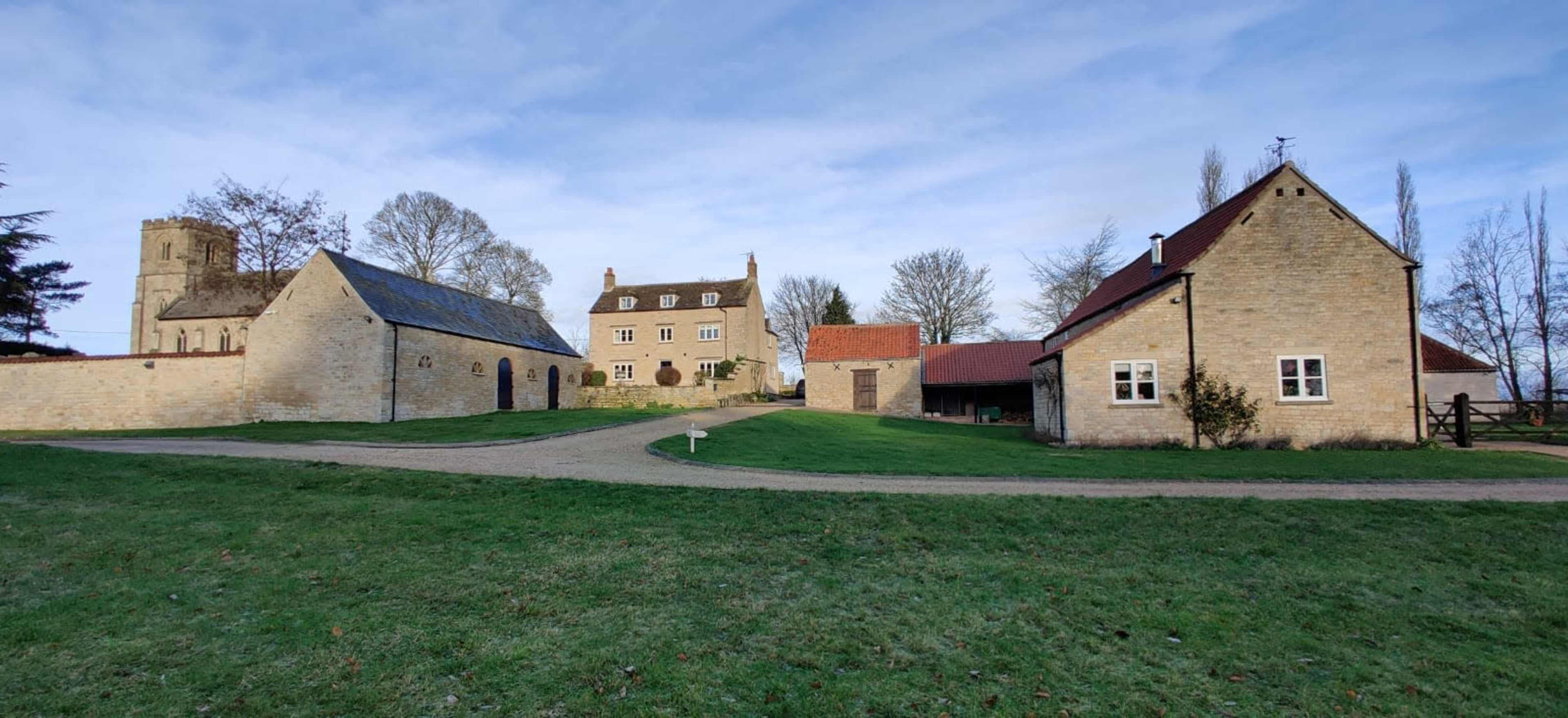 The image shows a rural landscape featuring multiple stone buildings, including a church and a large house, set against a backdrop of trees and a clear sky.