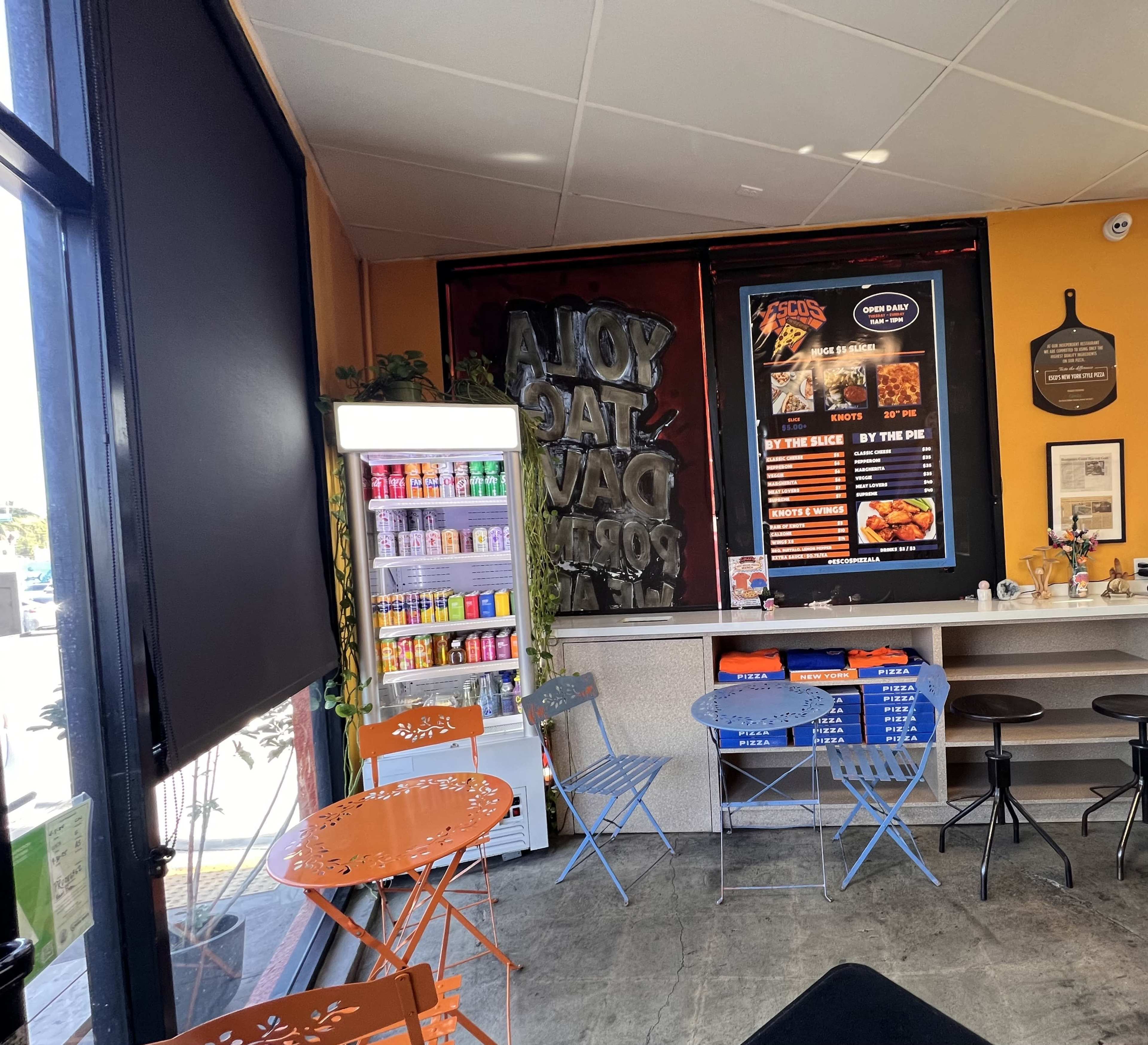 The image shows the interior of a casual eatery with colorful tables, a beverage cooler, and a menu displayed on the wall.