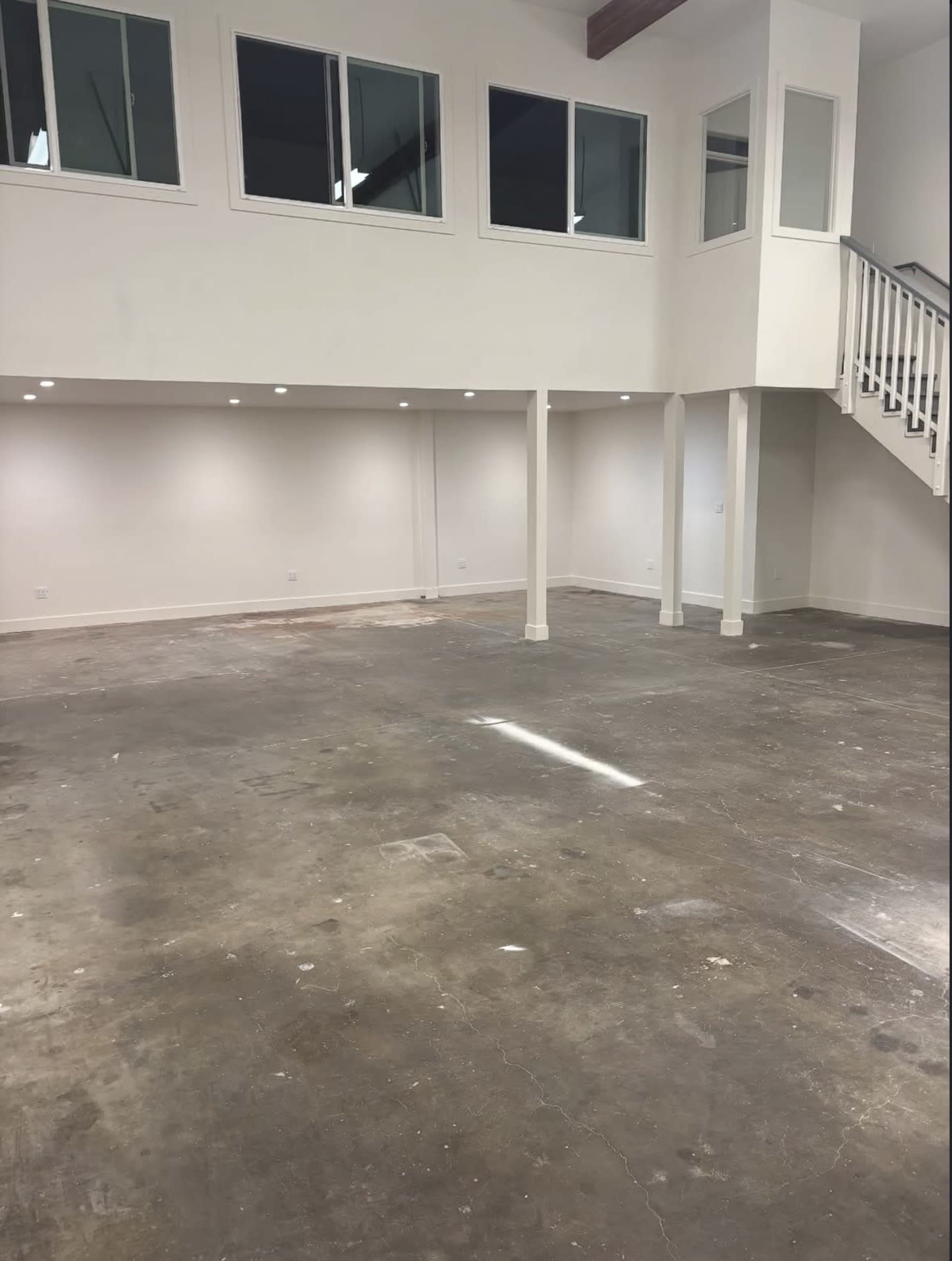 The image shows an empty interior space with a polished concrete floor, white walls, and large windows along one side, featuring a staircase leading to a raised area.