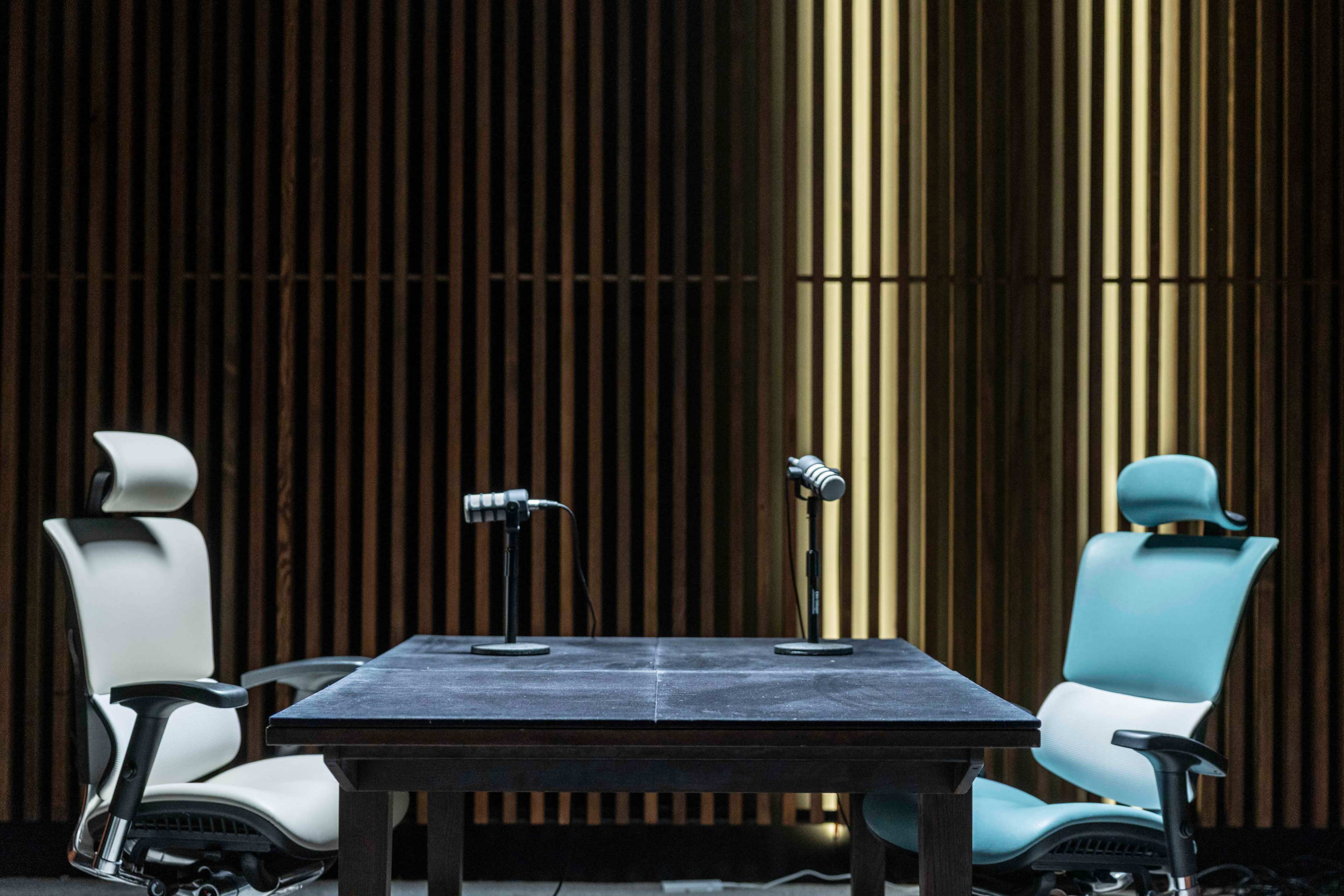 Two office chairs are positioned on either side of a dark wooden table, each equipped with a microphone, against a backdrop of wooden slats.