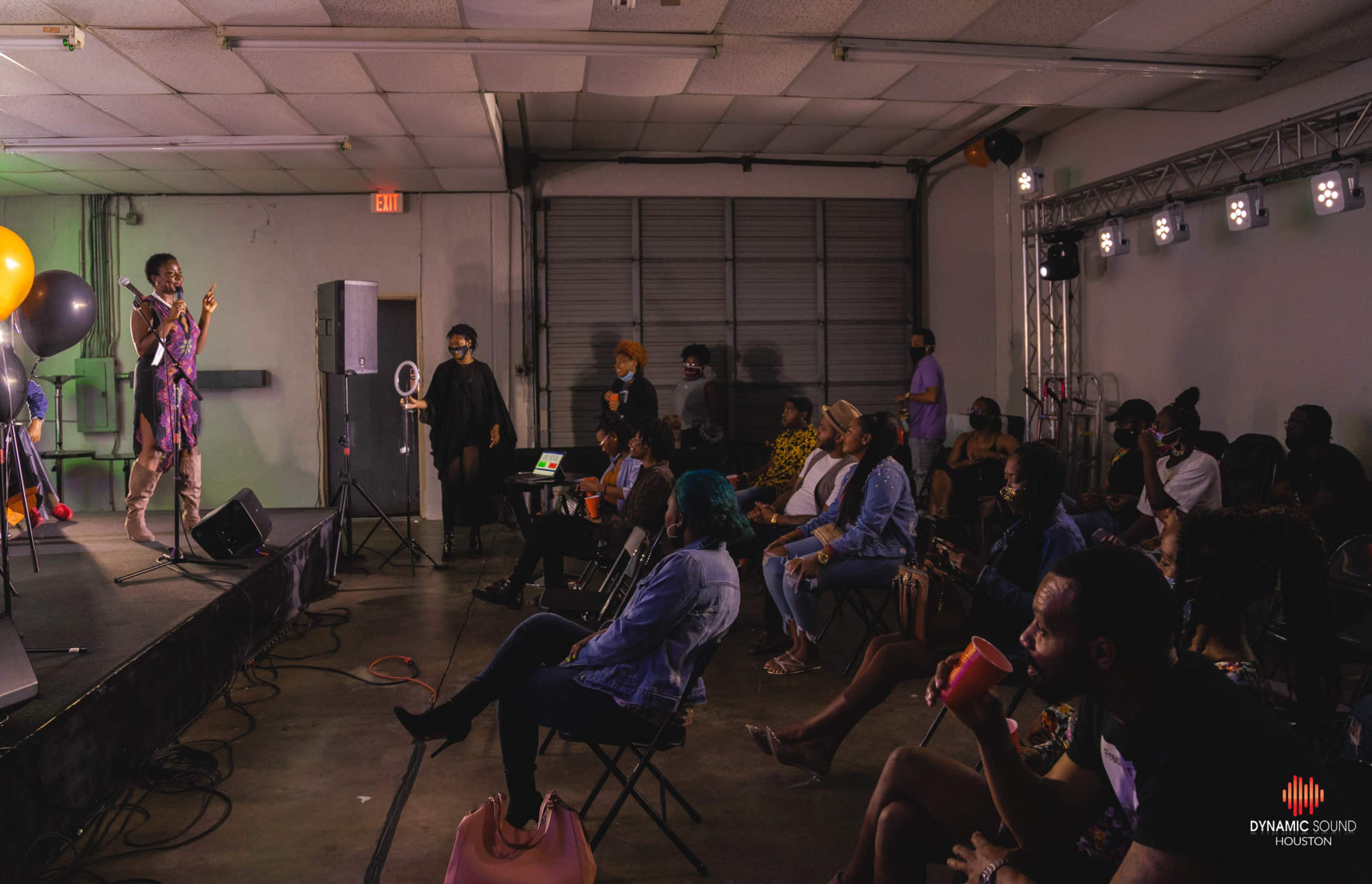 A performer stands on stage in front of an audience seated in folding chairs at an indoor event space.