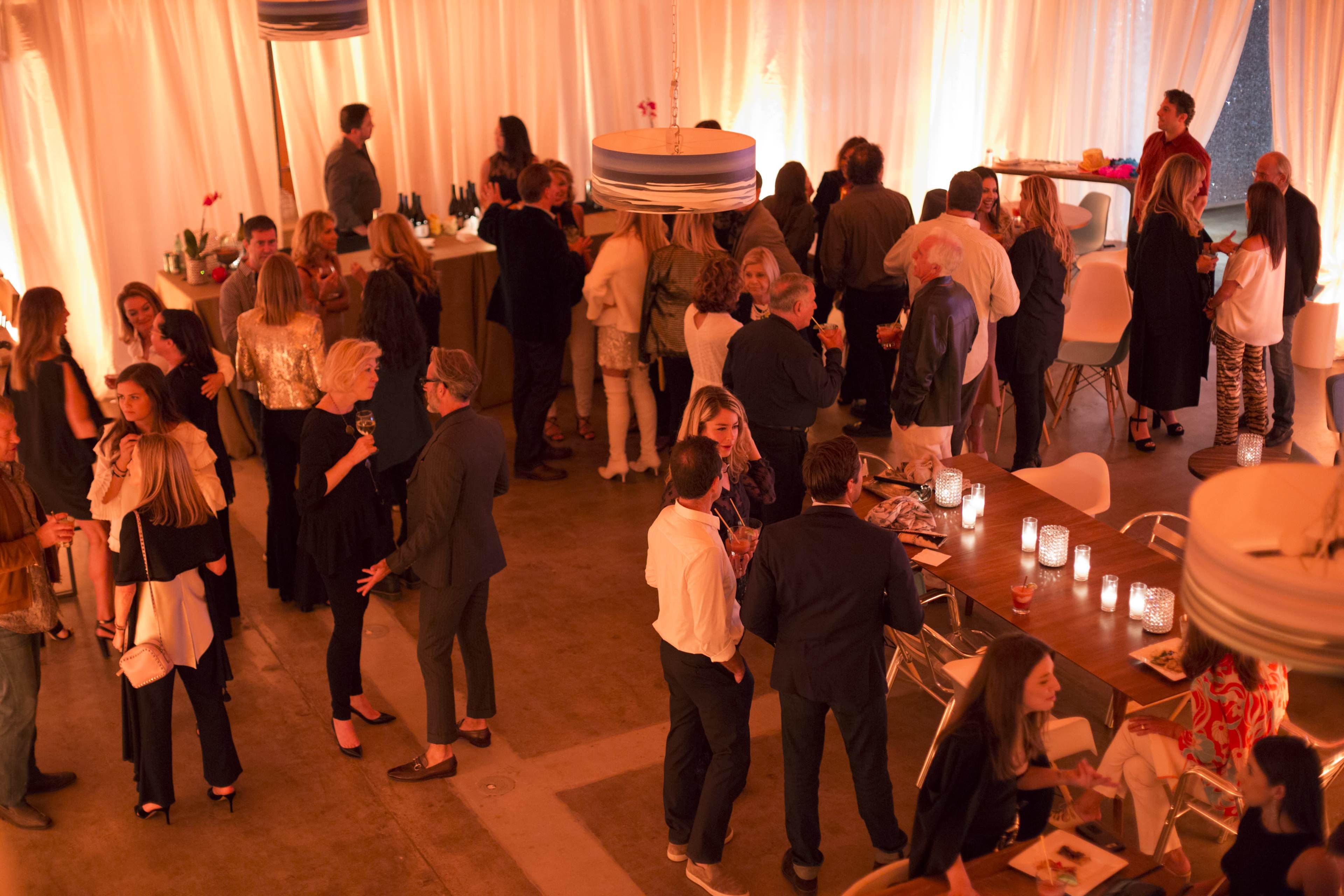 A social gathering is taking place in a warmly lit venue, with groups of people conversing and several tables set with drinks and snacks.
