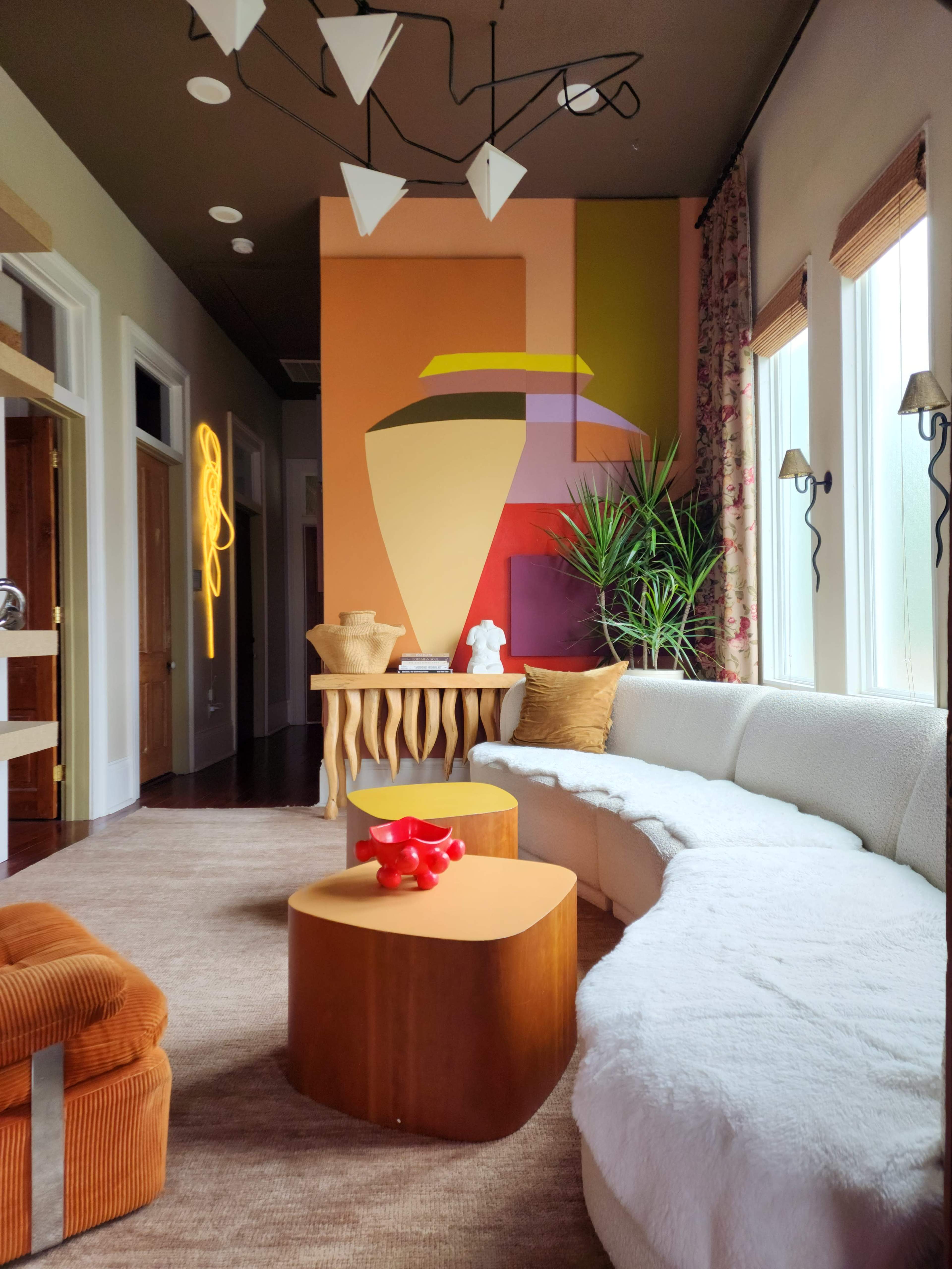 The image shows a modern living room featuring a curved white sofa, wooden accent tables, and colorful abstract wall art.